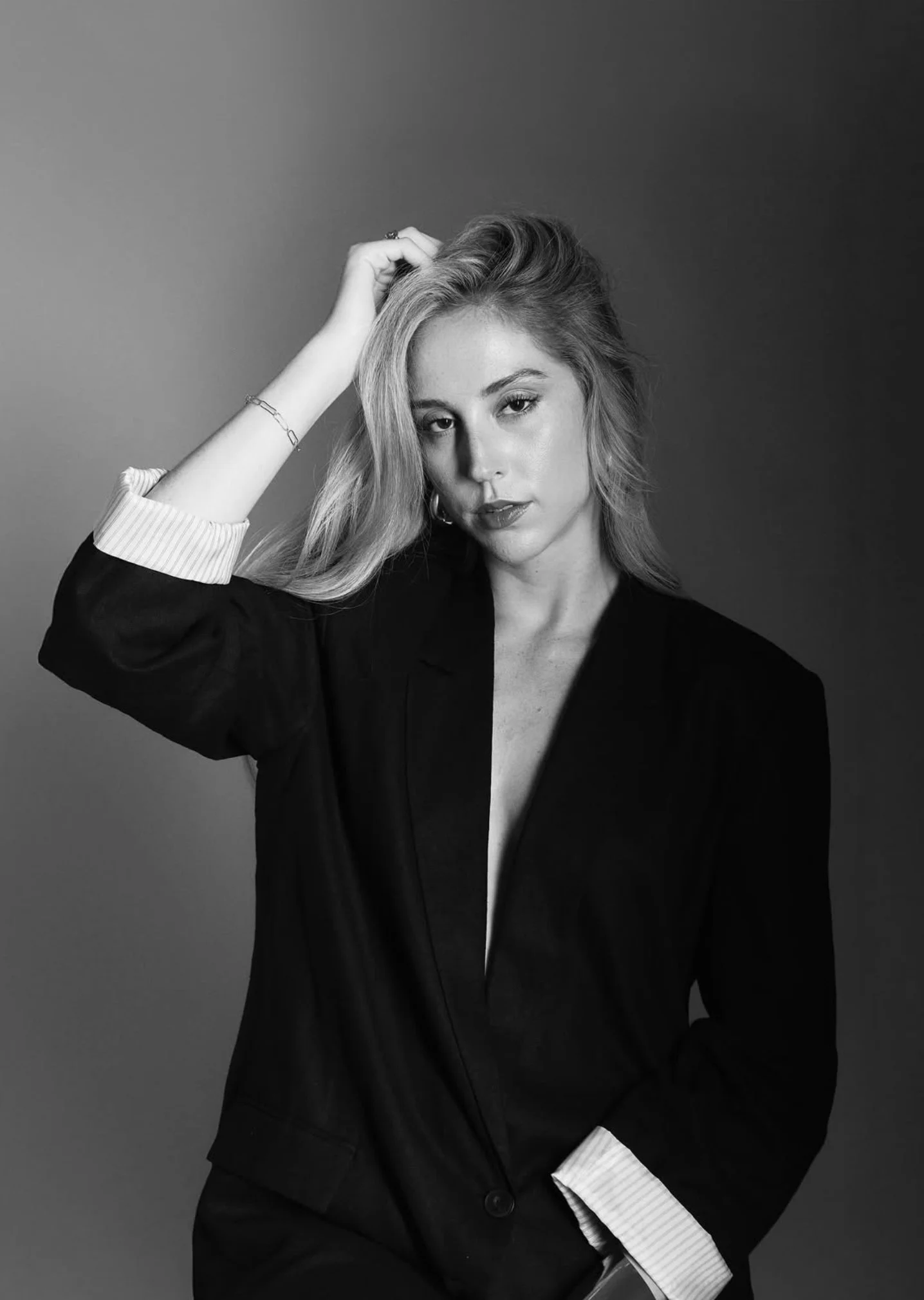 A woman with long hair, dressed in a blazer with a striped shirt underneath, posing against a gradient background, looking at the camera.