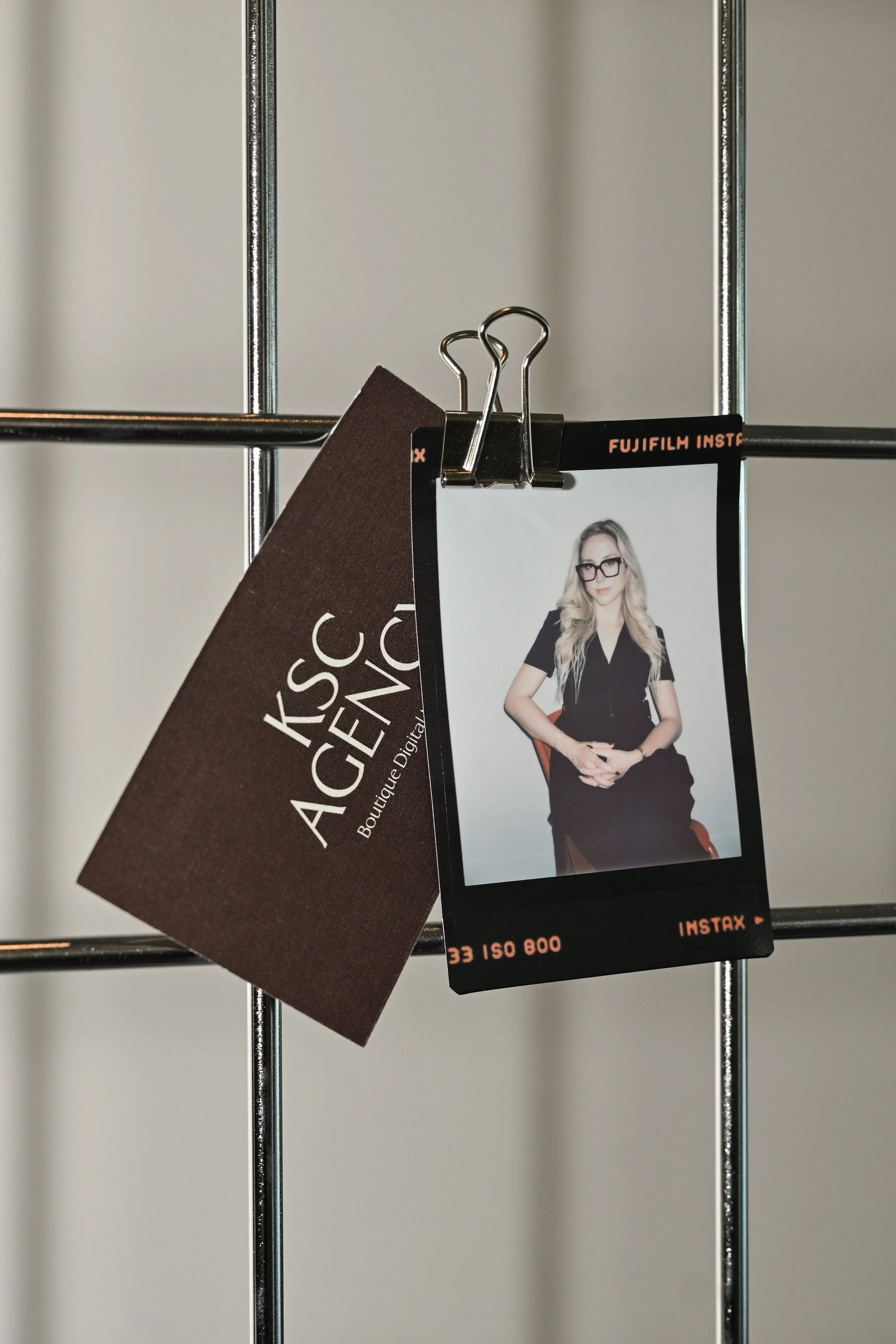A photograph of a photo and a business card hanging on a metal wire grid. The photo features a woman with long blonde hair, wearing glasses and a black outfit, sitting on an orange chair against a white background. The business card has the text "KISS ACCENT" and additional smaller text, attached to the grid with a black paperclip.