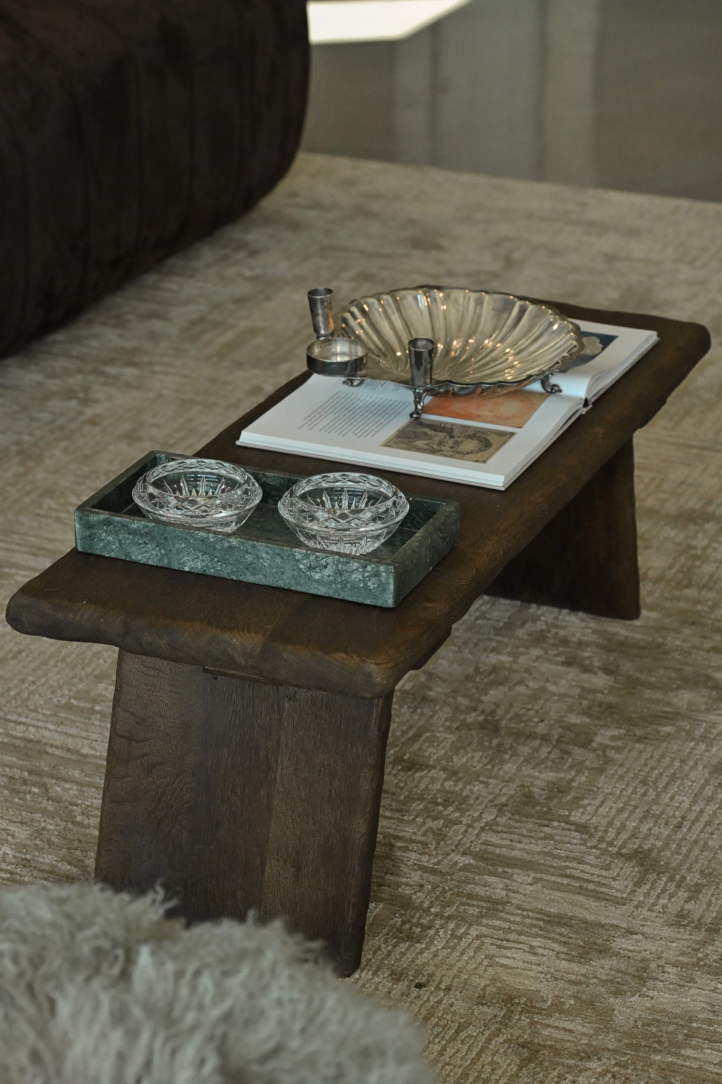 A wooden table with a silver shell-shaped dish, two glass bowls on a green tray, and an open magazine with images and text, on a beige carpeted floor.