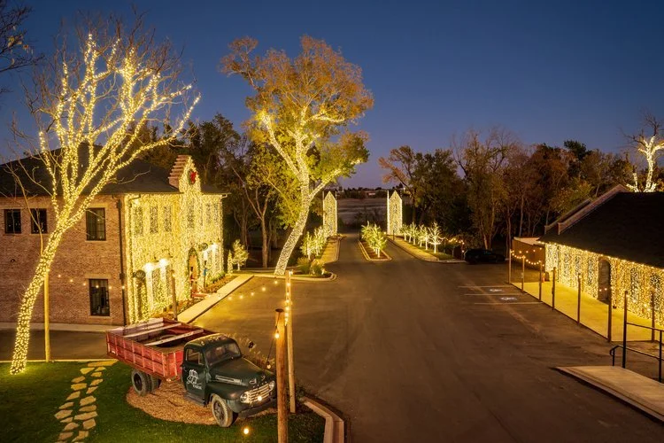 A street decorated with Christmas lights on trees and buildings, with a truck parked in the foreground, at dusk or night.