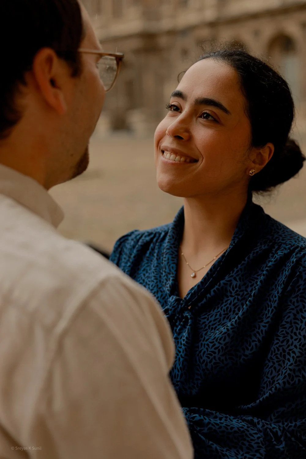 A woman smiling warmly at a man in an outdoor setting with historic architecture in the background.