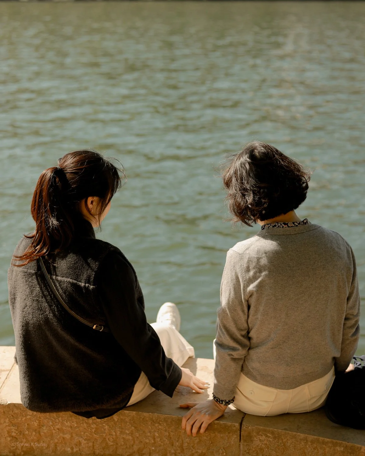 Two women sitting on a stone ledge by a body of water, facing away from the camera, engaged in conversation.