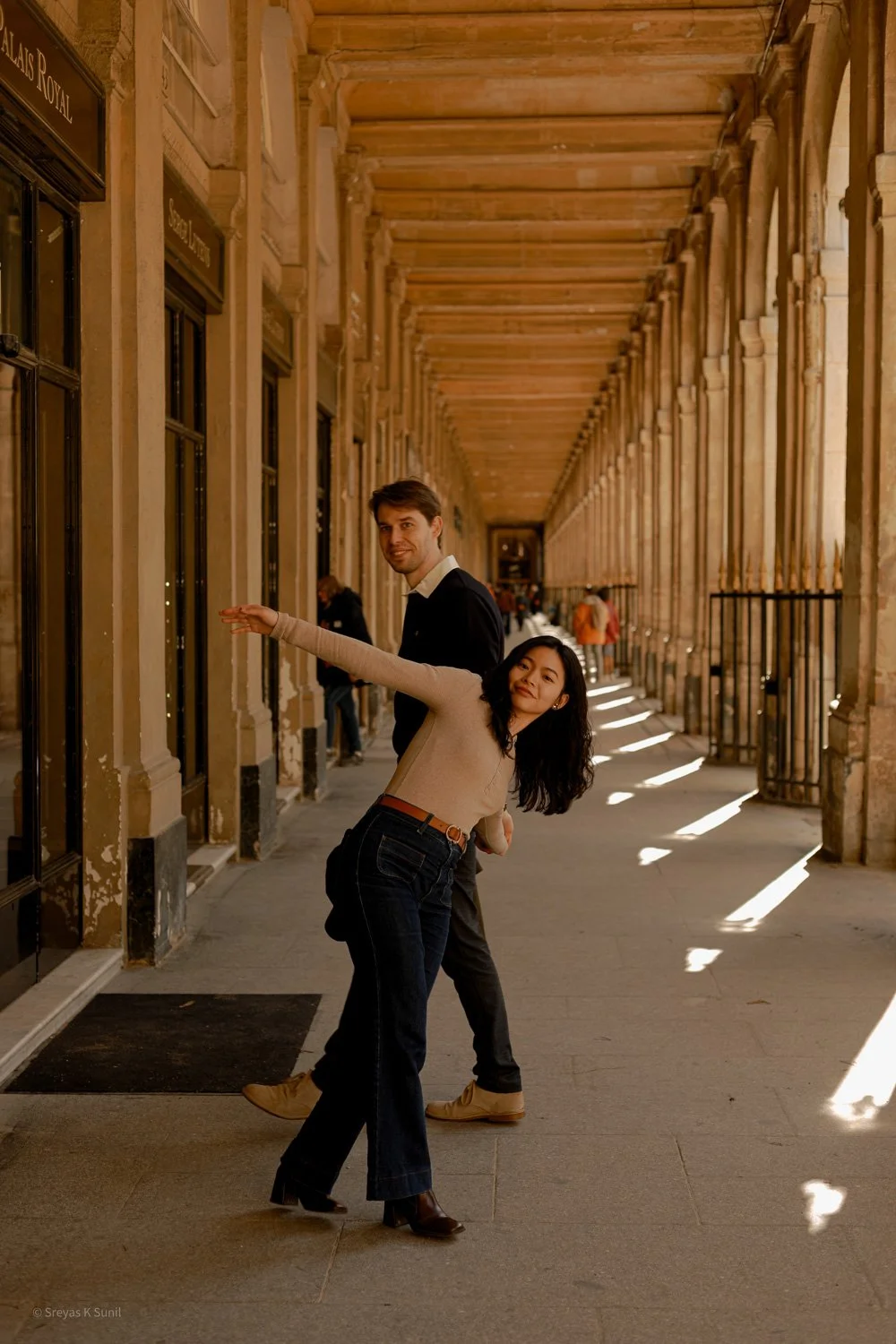 A couple dancing in a covered walkway with columns, sunlight creating striped shadows on the ground, in a historical or architectural setting. Palais Royale