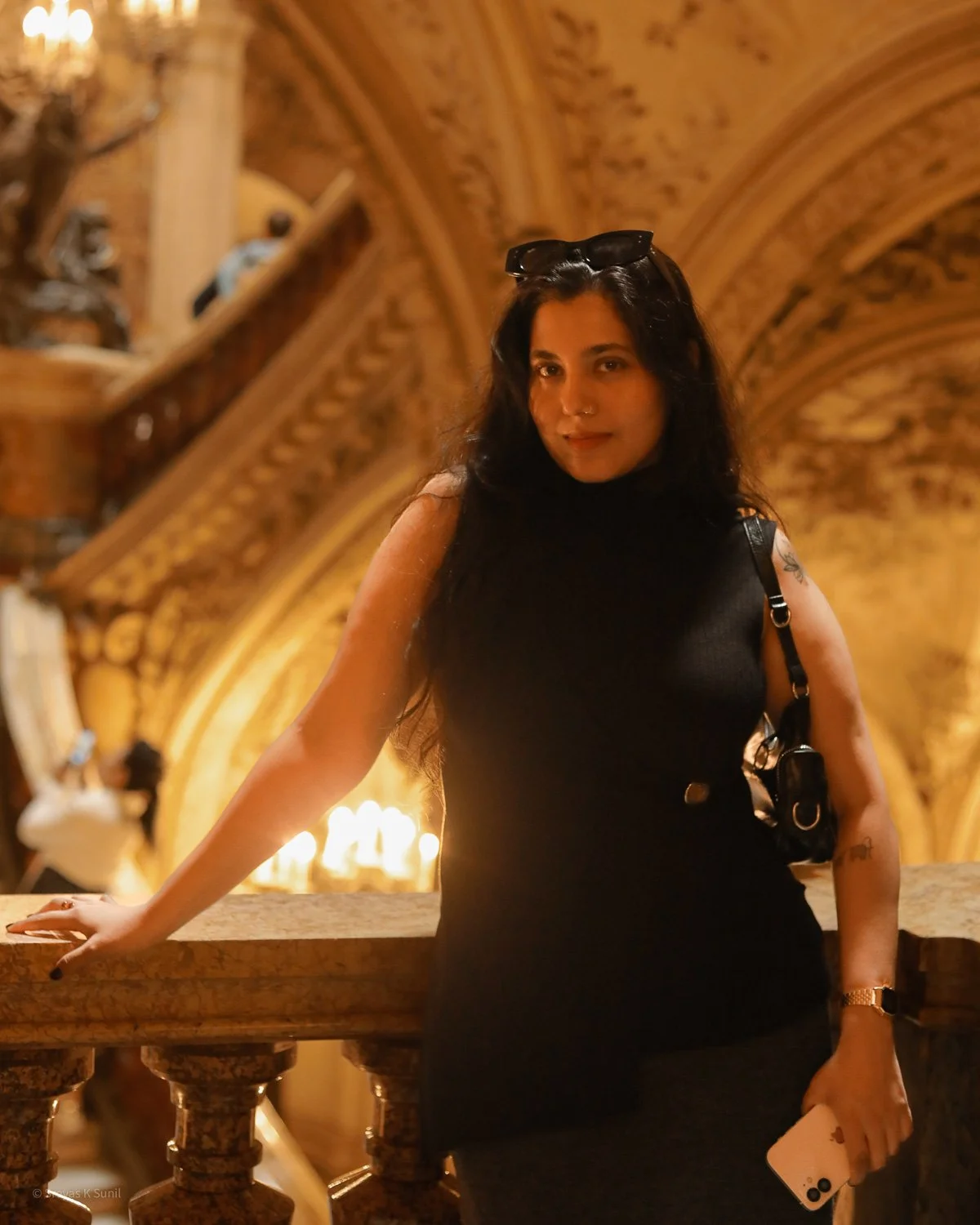 A woman with long dark hair wearing a black sleeveless dress, sunglasses on her head, posing on a marble railing inside an ornate, dimly lit historic building with intricate woodwork and chandeliers.