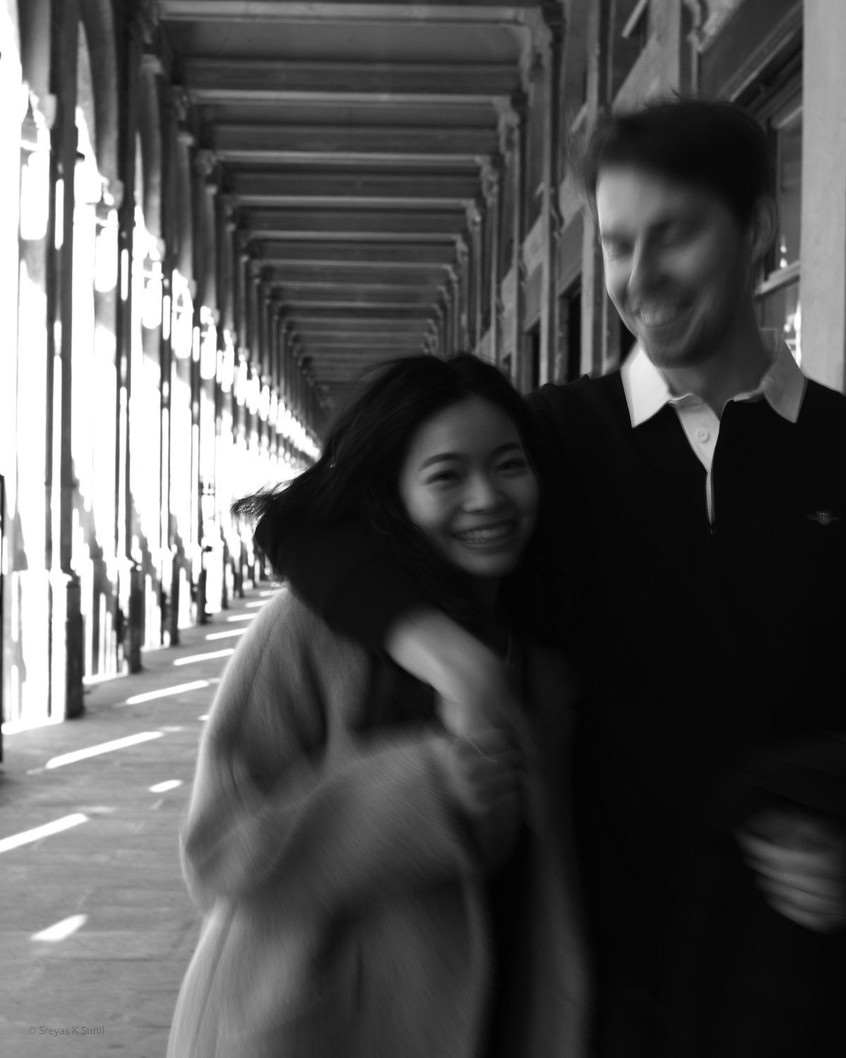 Happy couple hugging each other in a black-and-white photo, standing on a covered outdoor walkway with a patterned ceiling and columns on the side. Palais Royale