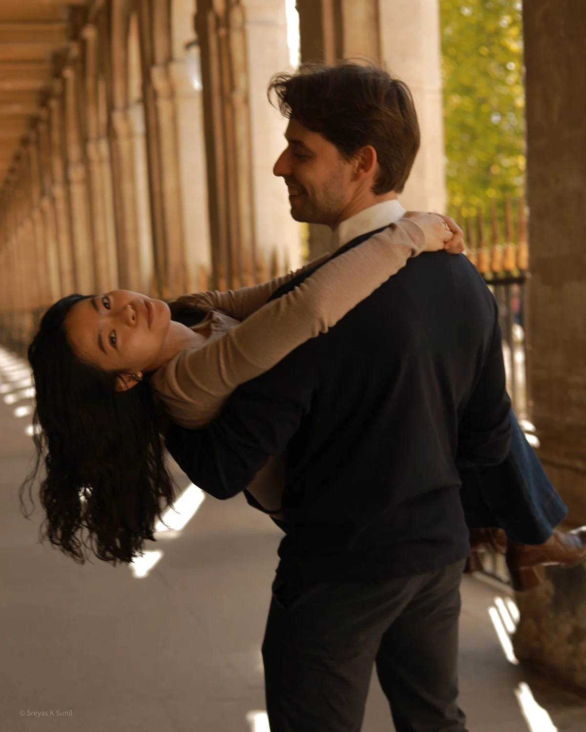 A man in a dark suit holding and dancing with a woman with long dark hair and tan clothing in a covered outdoor walkway.