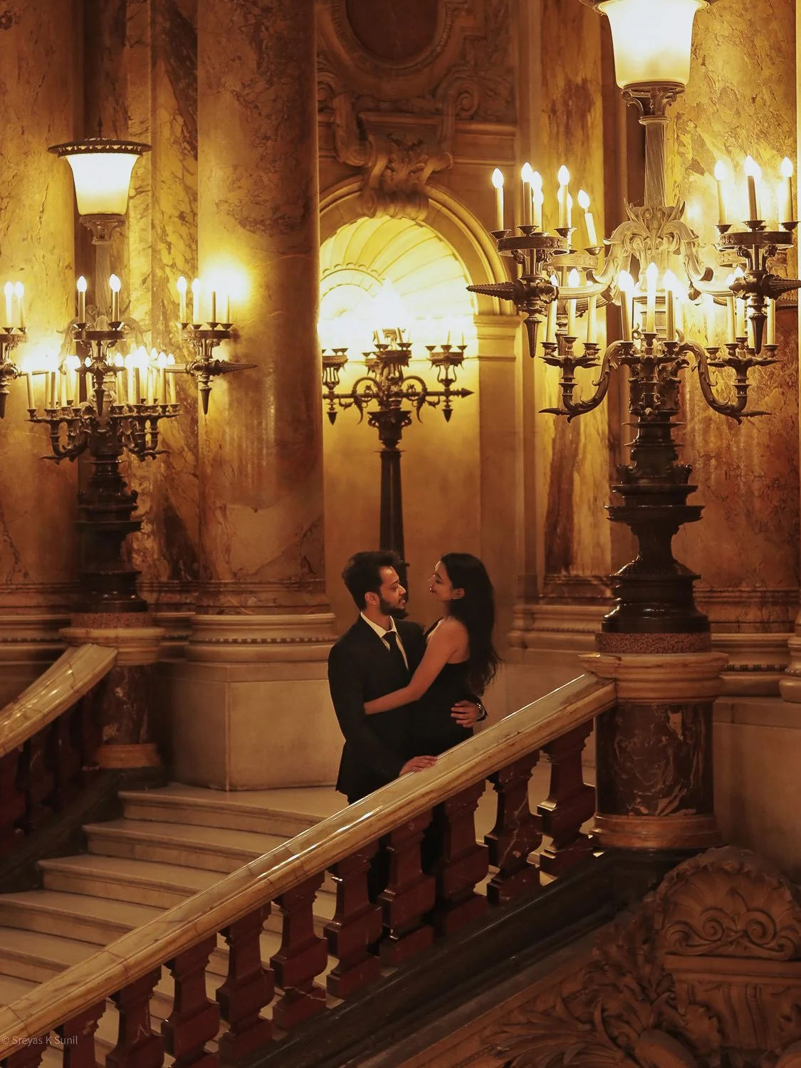 A romantic couple dressed in formal attire embracing on a grand staircase inside a luxurious, ornate hall with marble columns, chandeliers, and warm yellow lighting. Paris Opera