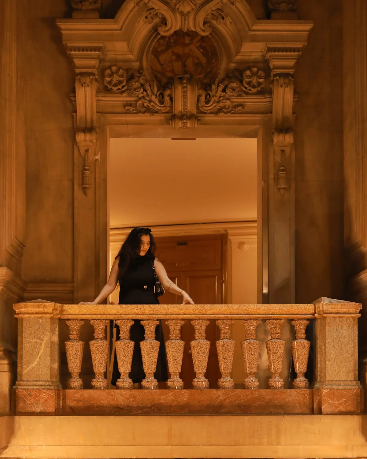 Woman with dark hair wearing black dress, standing on a marble balcony inside an ornate, wood-paneled room with elaborate carvings and decorative molding.