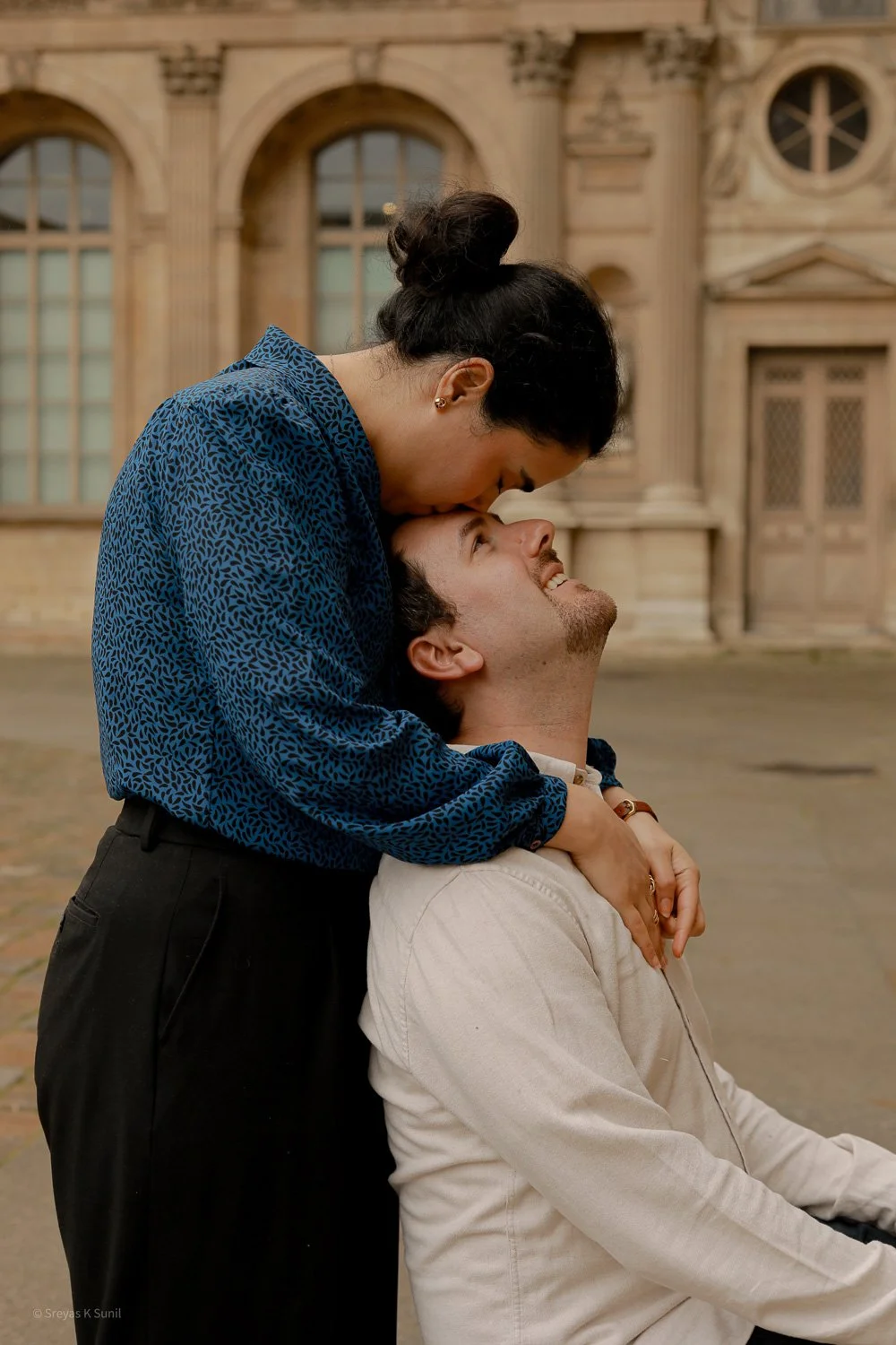 A woman leans over to kiss a smiling man sitting in front of her outdoors in front of a historic building.