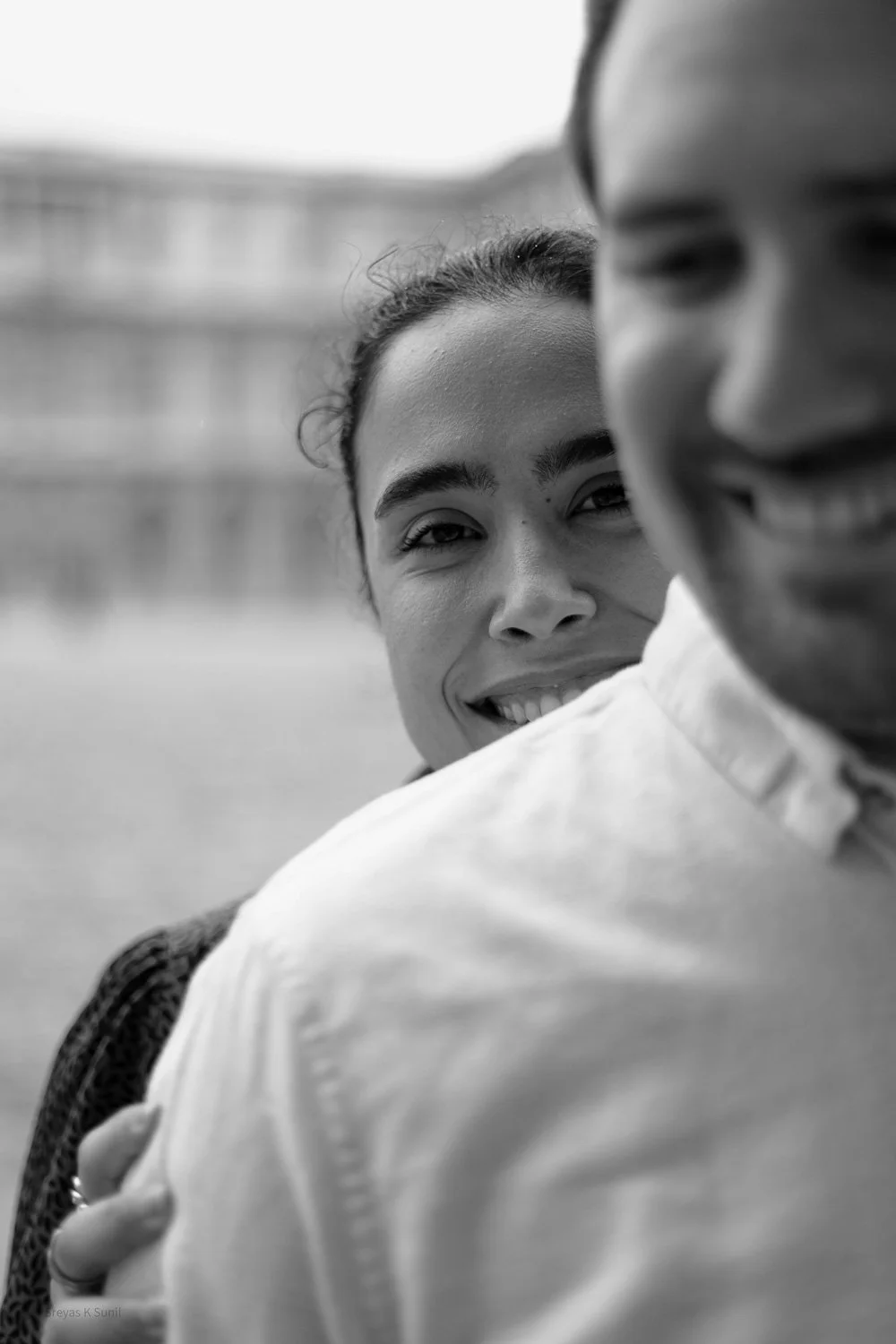 A black-and-white photo of a woman smiling at the camera, partially obscured by her man's shoulder, who is also smiling.