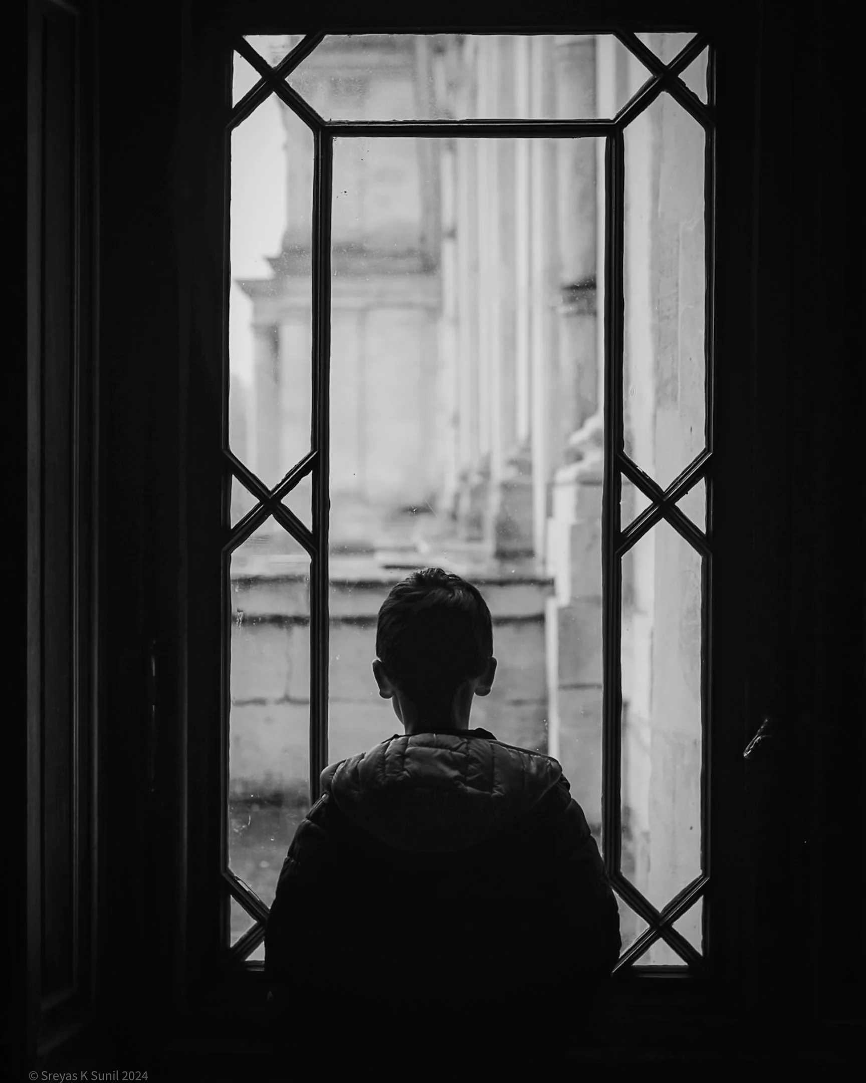 Silhouette of a child looking out a window, black and white photo, city building visible outside.