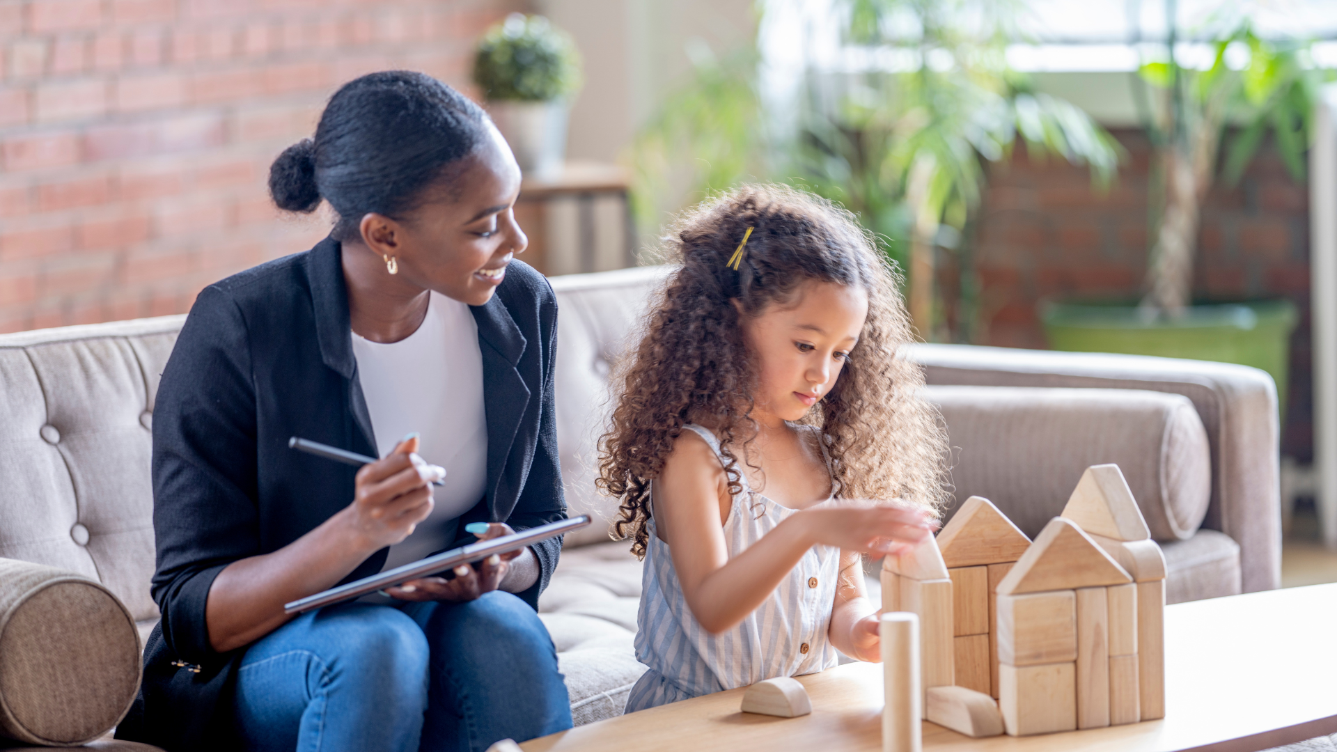 A woman and a young girl sitting on a sofa, engaging with wooden building blocks, with the woman holding a tablet and smiling at the girl, in a bright room with green plants and a brick wall in the background.