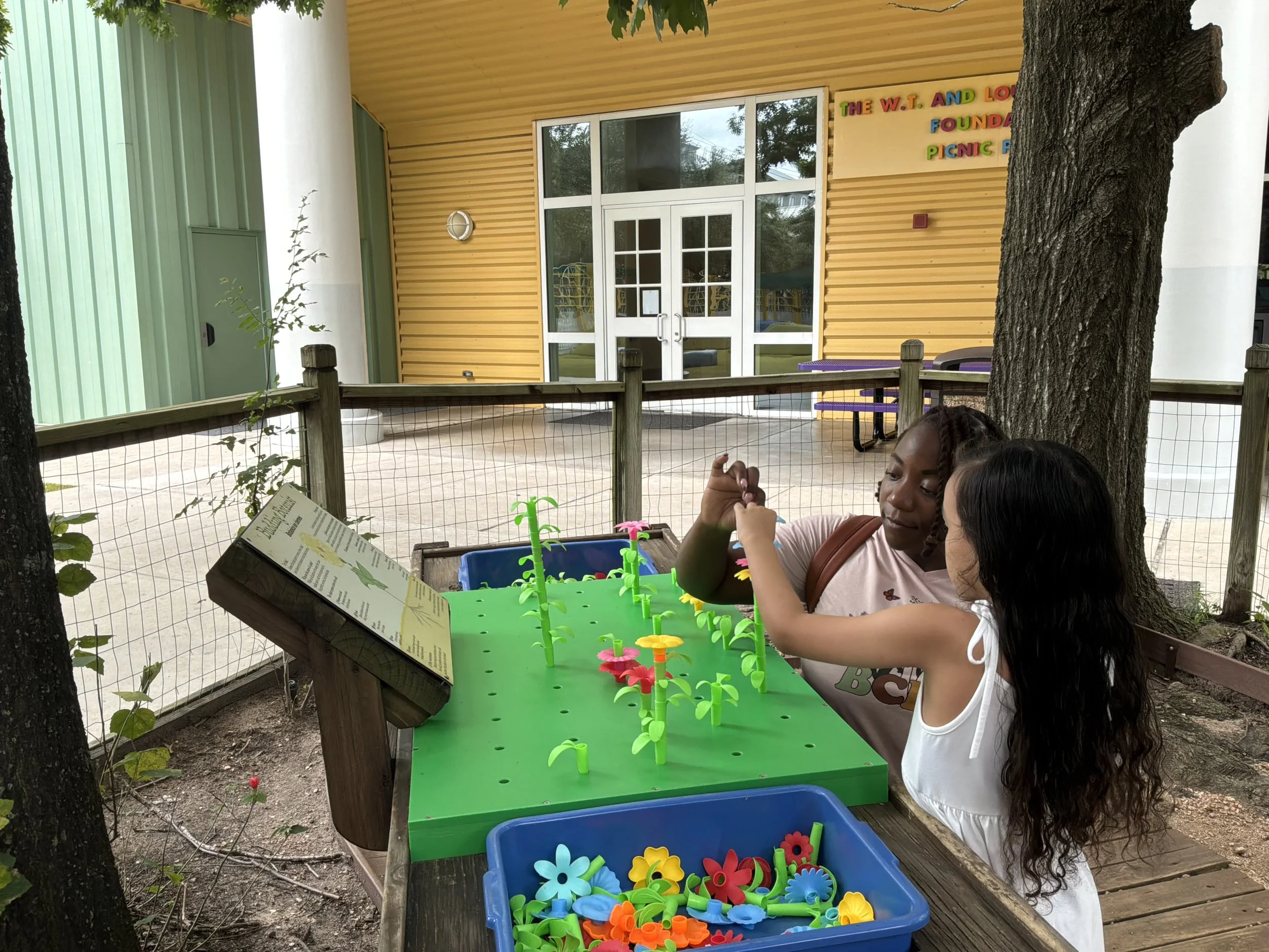 A female therapist in red scrubs giving a high five to a young girl sitting on a padded gym block in a physical therapy or occupational therapy session at a therapy gym with colorful padded mats and equipment.