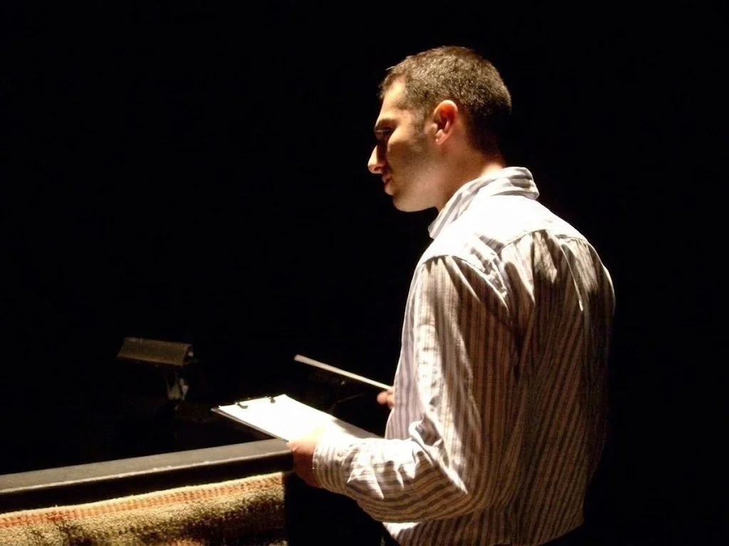A man in a striped shirt holds sheet music and stands near a piano, illuminated by a spotlight against a dark background.