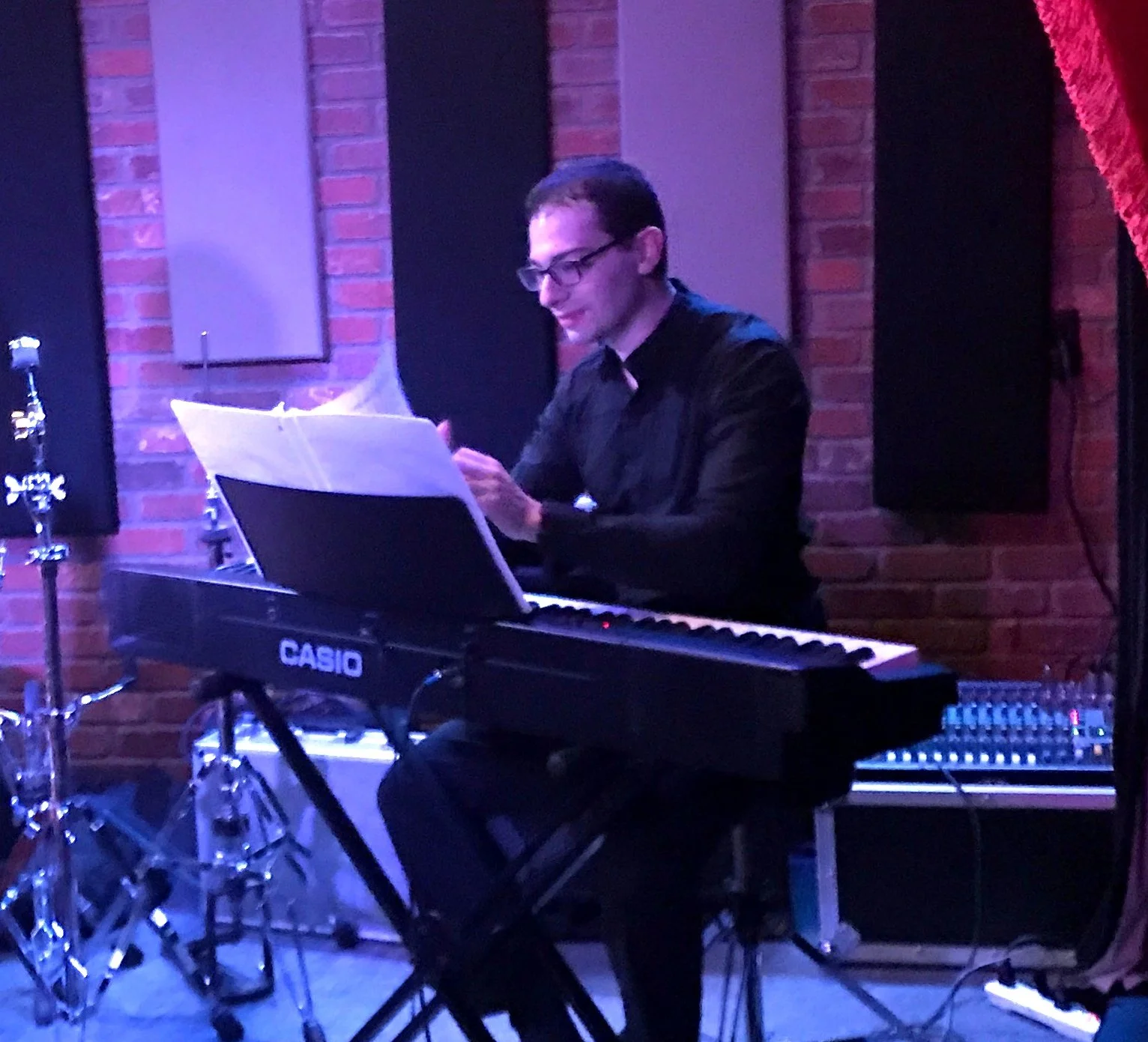 A man in glasses and a black shirt playing a Casio keyboard on stage with a brick wall background, music stand with sheet music, and a red curtain on the side.