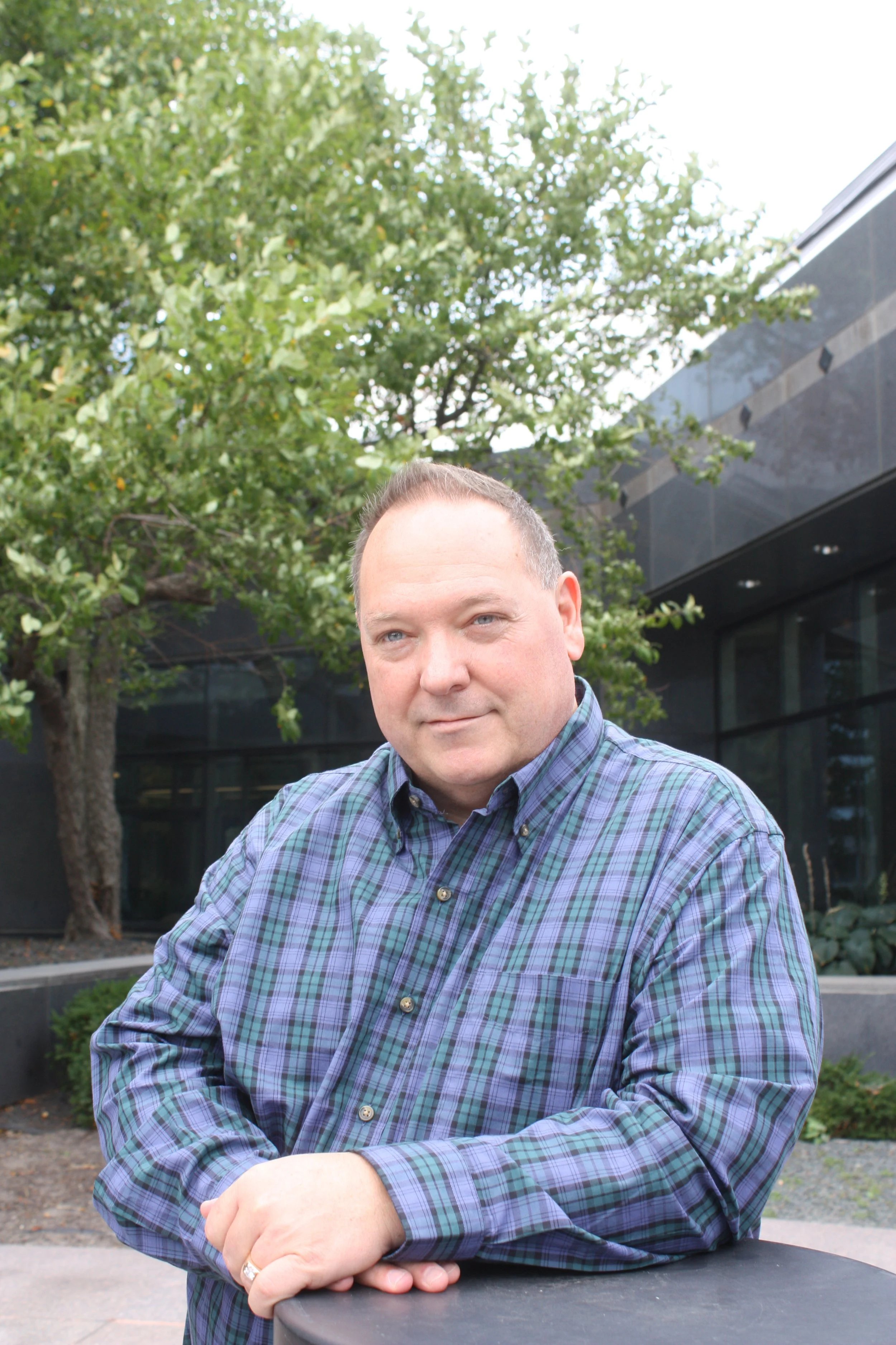 A man in a plaid shirt sitting outdoors with trees and a modern building in the background.