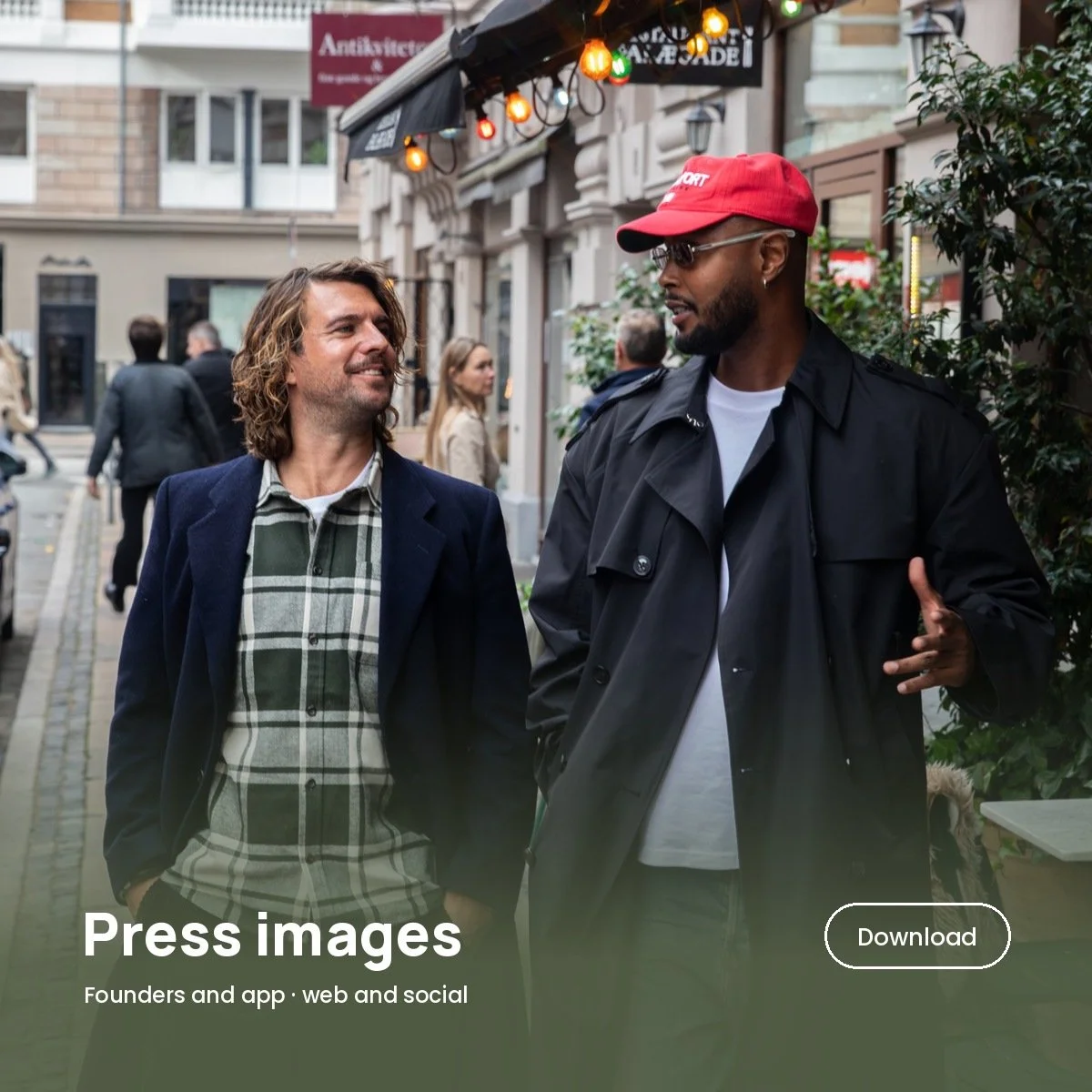 Two men walking and talking on a city sidewalk with shops and pedestrians in the background. One has curly hair, a beard, and is wearing a blazer over a plaid shirt. The other is wearing sunglasses, a red cap, and a black jacket, gesturing with one h