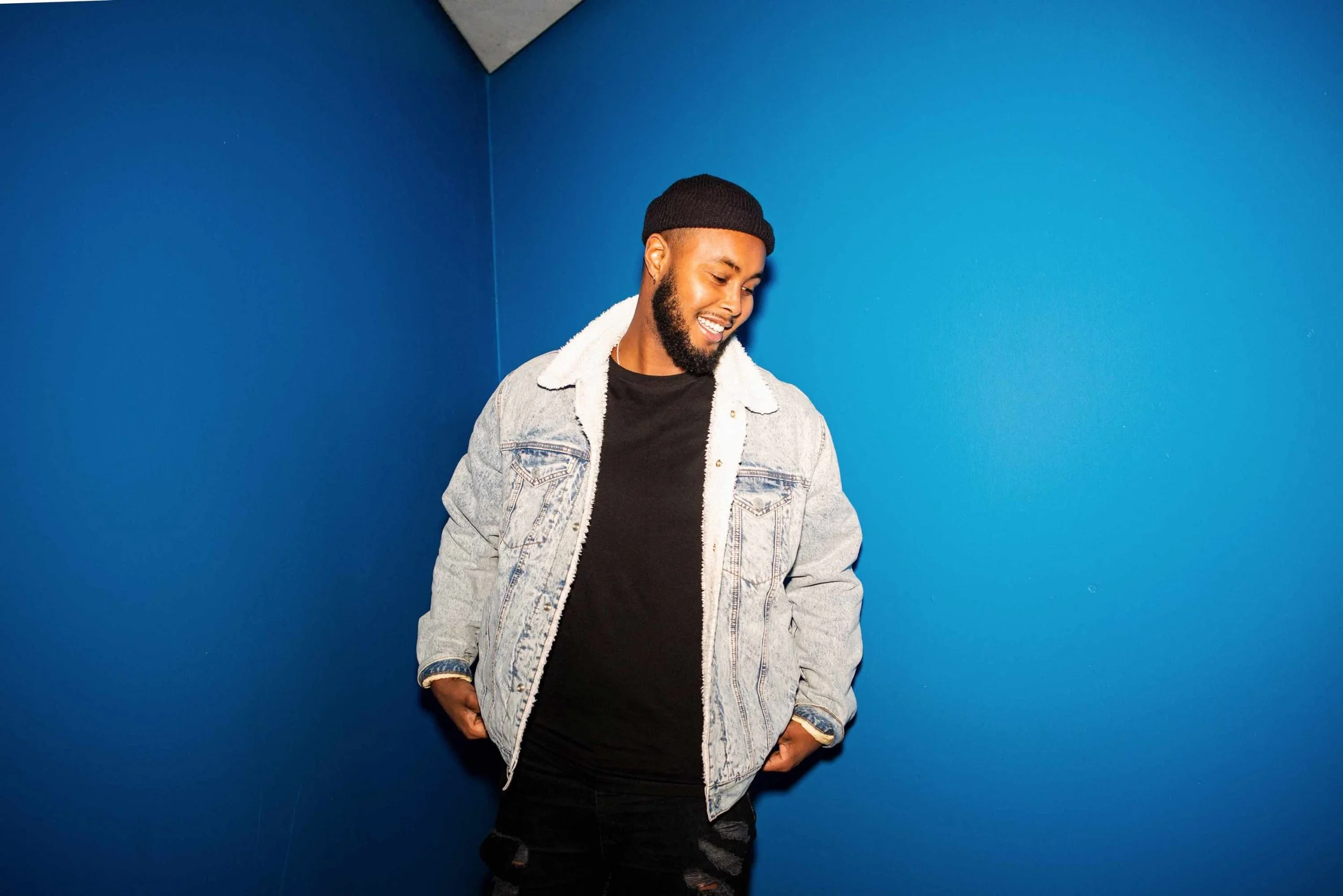 A young man with dark skin and a beard wearing a black beanie, denim jacket with a shearling collar, and black pants, standing against a blue wall, smiling and looking down.