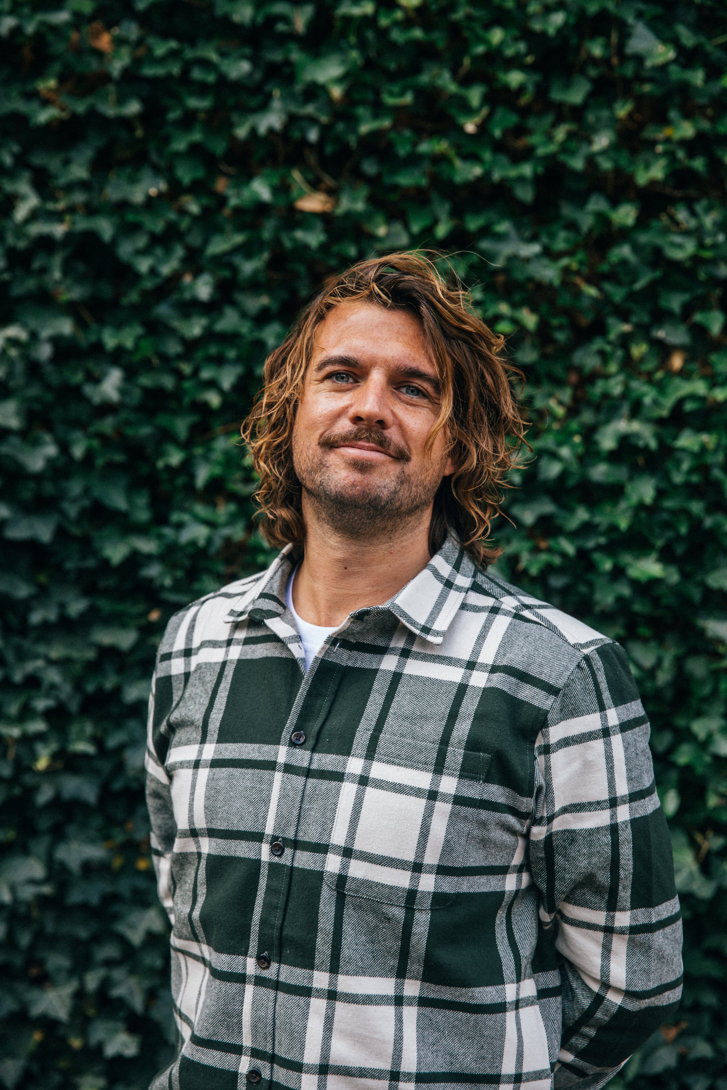 A man with wavy, shoulder-length blonde hair and a light beard, wearing a black and white checkered shirt, standing outdoors in front of a dense green ivy background.