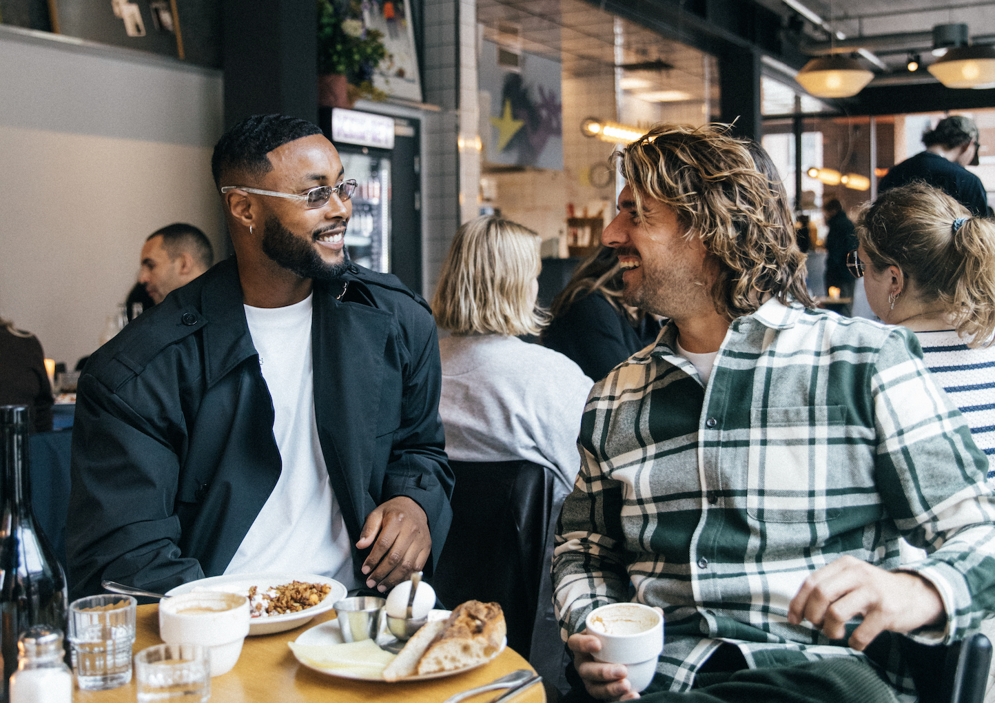 Two men are sitting at a restaurant table, smiling and engaging in conversation. One man has dark skin, short hair, glasses, and is wearing a black jacket and white t-shirt. The other man has lighter skin, long curly hair, and is wearing a green plaid shirt. On the table are plates of food, including rice and bread, along with a cup of coffee or tea. Other diners are visible in the background.
