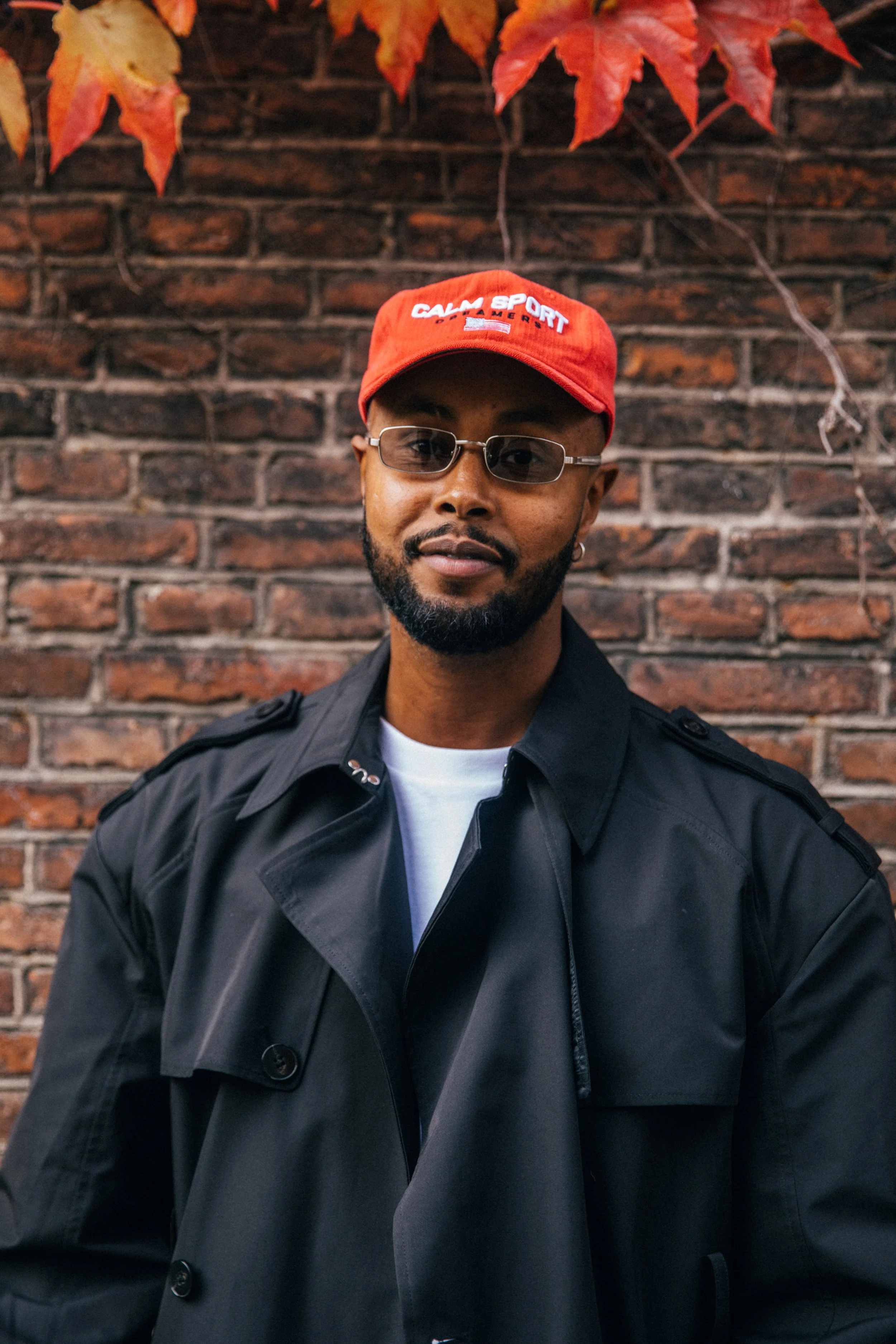 A man wearing a red cap, glasses, and a black trench coat stands in front of a brick wall with red and orange fall leaves.
