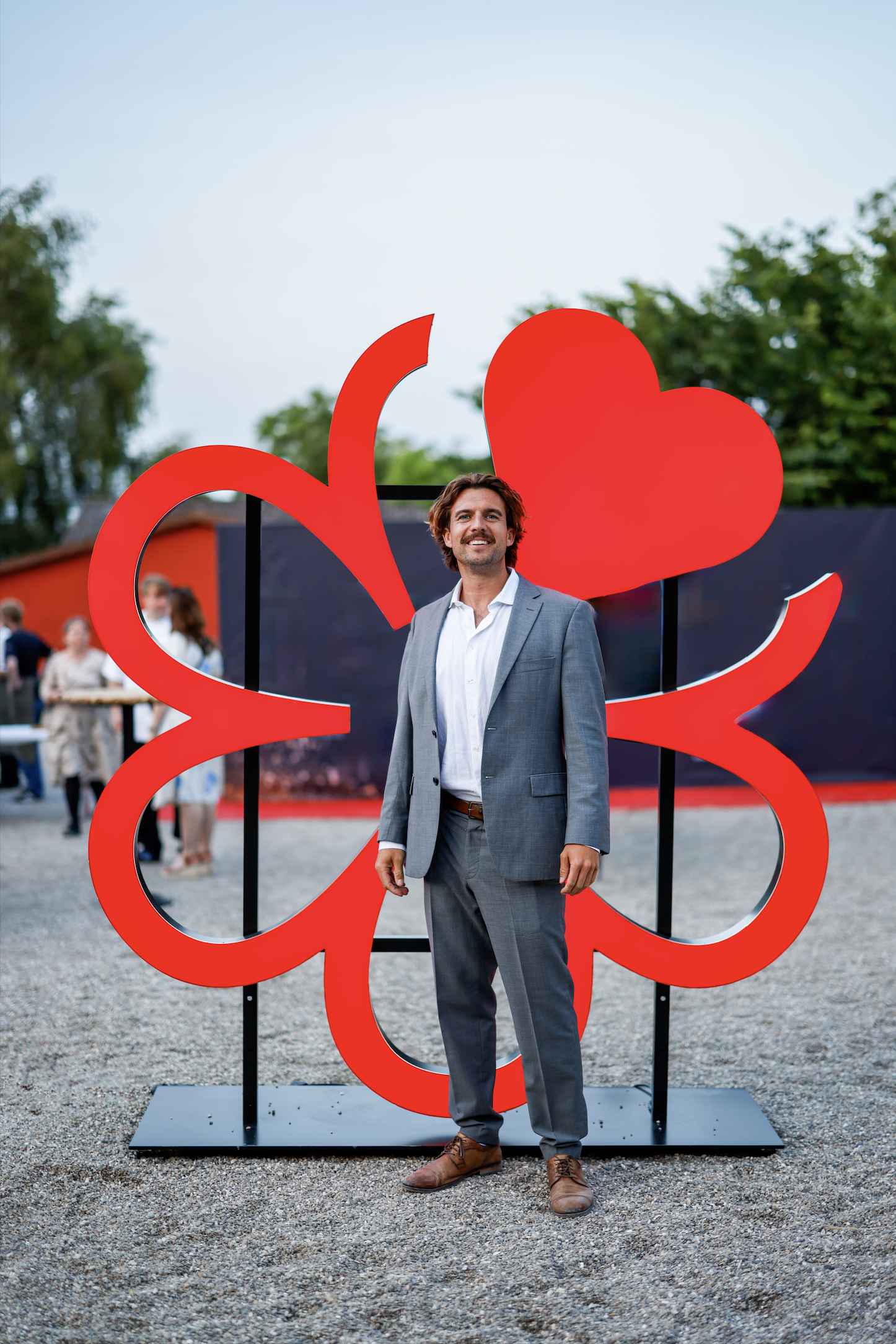 A man in a gray suit standing outdoors in front of a large red clover-shaped sculpture, smiling at the camera.
