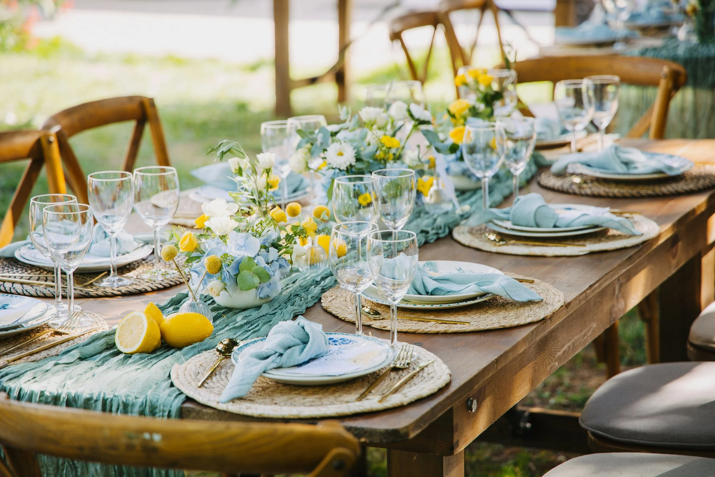 Outdoor dining table decorated with a green table runner, floral centerpiece with white and yellow flowers, plates, wine glasses, napkins, and lemons, set for a meal in a garden or backyard