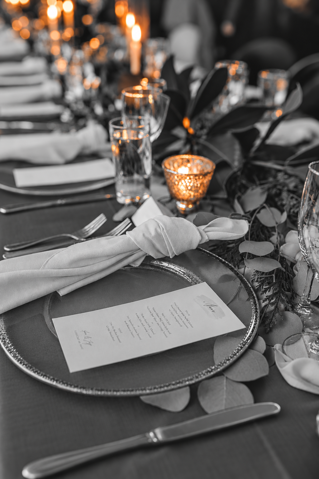 Elegant table setting with a menu, silverware, wine glass, and candlelight, decorated with greenery for a formal event.