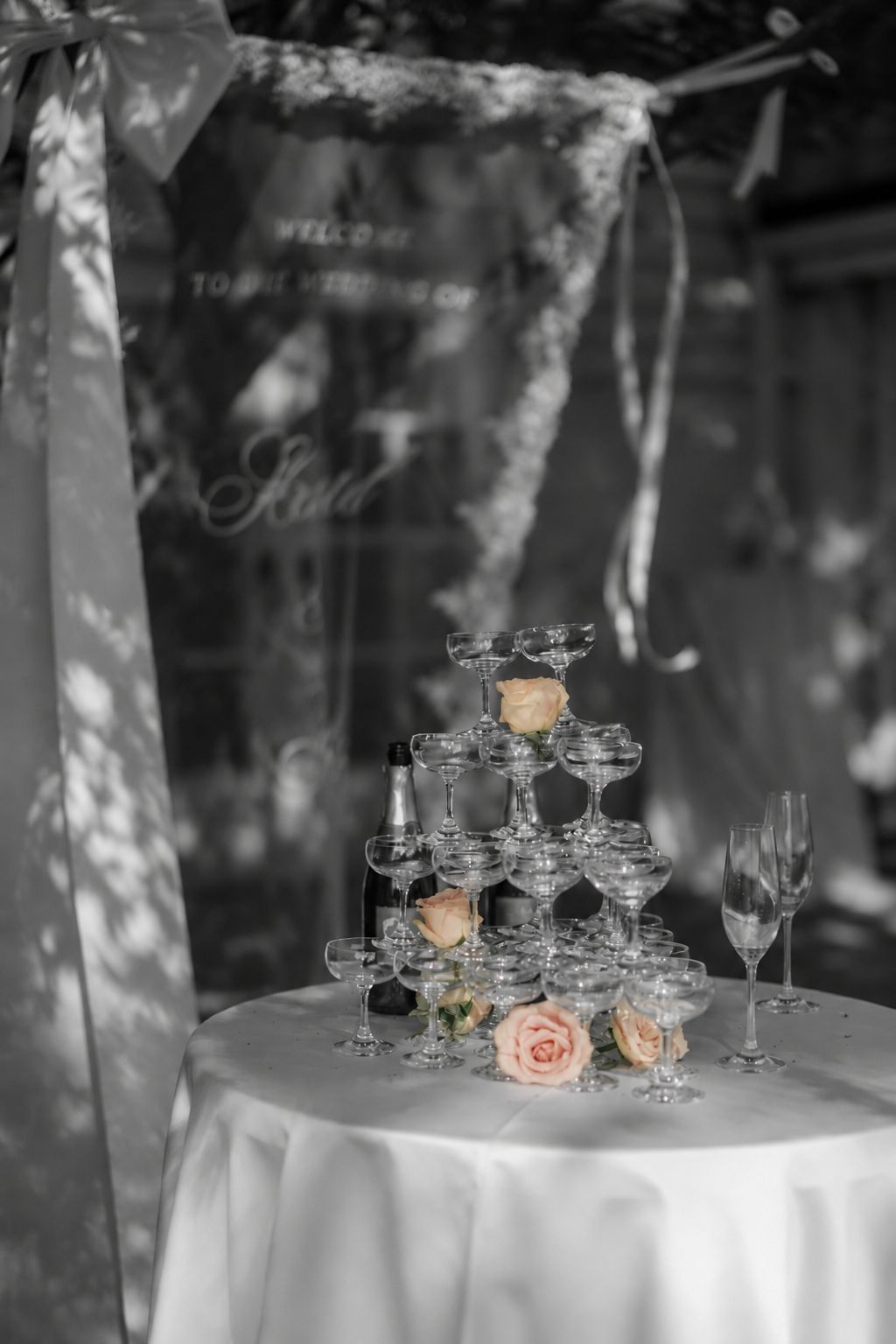 A champagne tower decorated with pink roses on a white tablecloth, with champagne bottles and glasses nearby, likely set for a celebration or wedding reception.