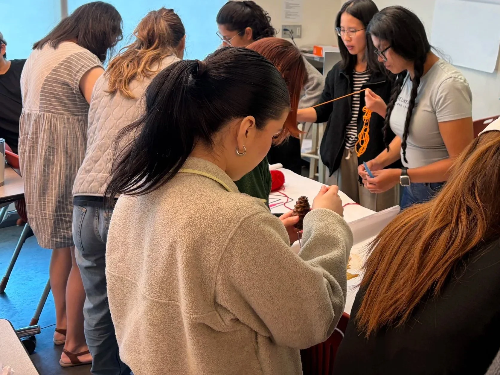 A group of women gathered around a table, engaging in a craft activity related to knitting or crocheting, with some holding yarn and needles, in a classroom or workshop setting.