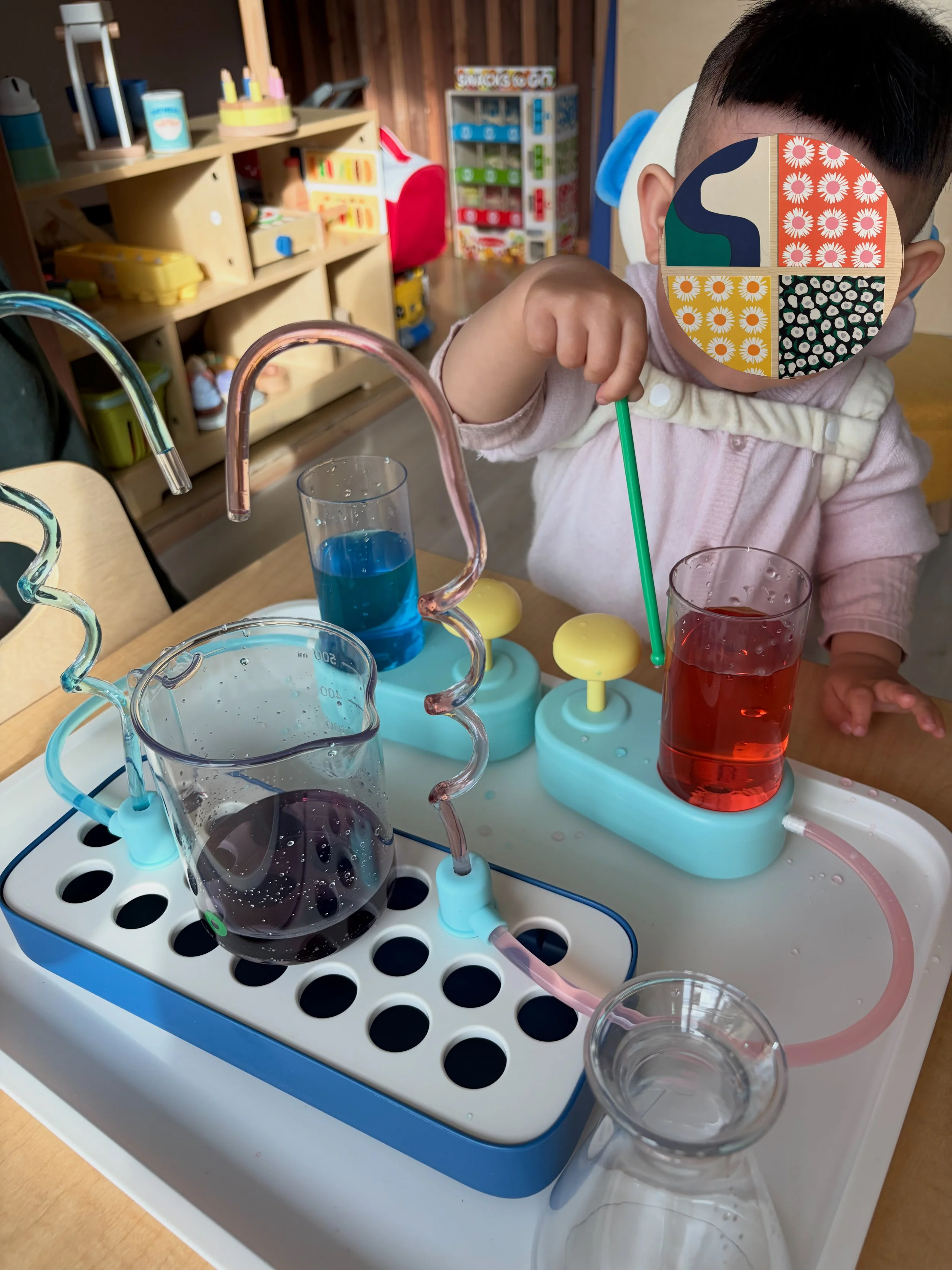 Child playing with colorful science experiment setup including beakers with blue and red liquids, tubes, and a tray, in a playroom or classroom setting.