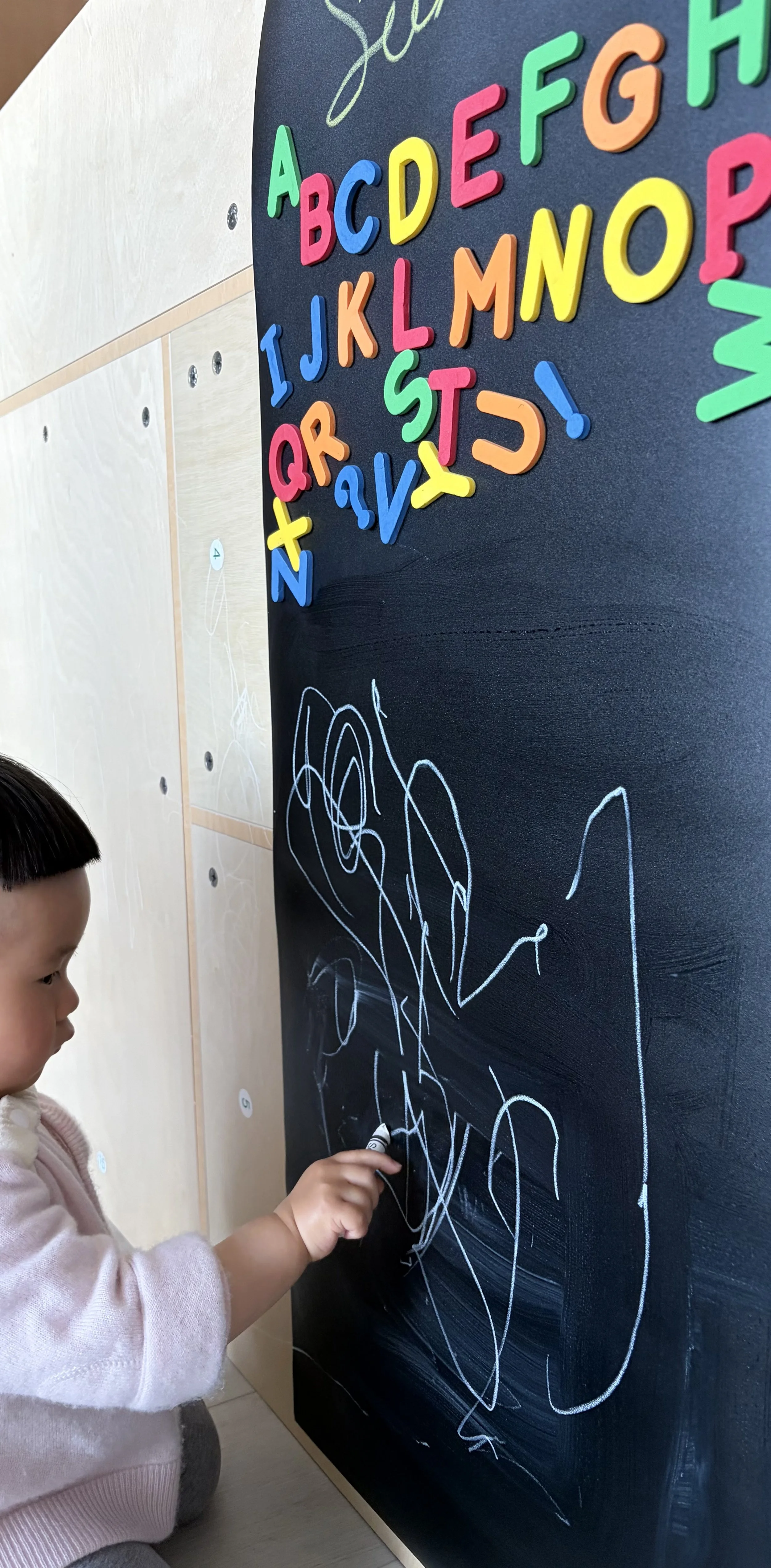 A young girl is drawing with chalk on a black chalkboard, creating scribbles. Colorful foam alphabet letters are attached to the top of the chalkboard, spelling out the alphabet from A to P.