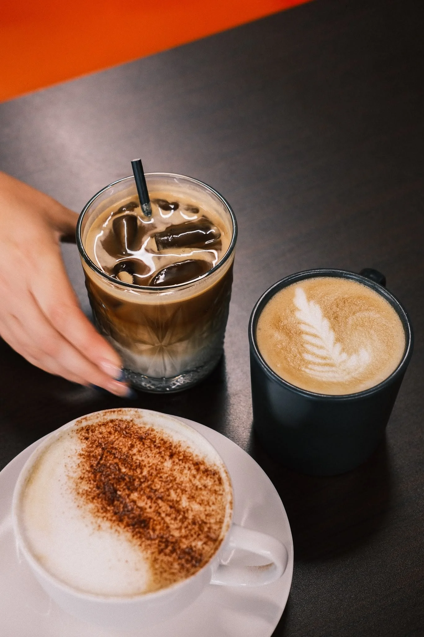 A hand holding a glass of iced coffee with a black straw, next to a latte in a black cup with foam art, and a cappuccino topped with cinnamon in a white cup, on a dark table.