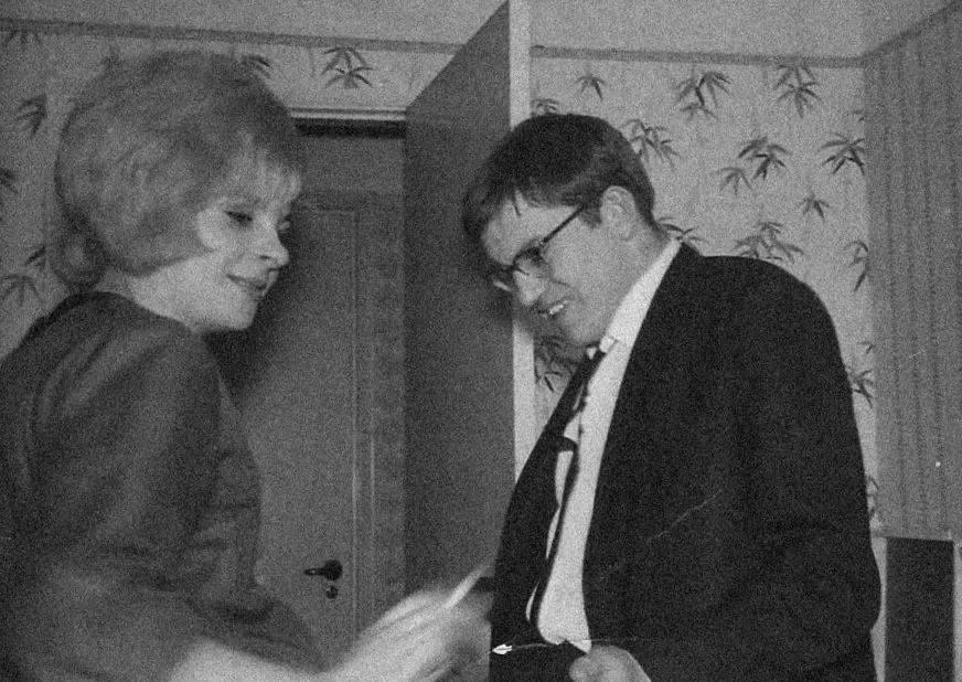 Two young men laughing and smiling at each other indoors, one wearing glasses and a suit, the other with longer hair and a casual jacket.