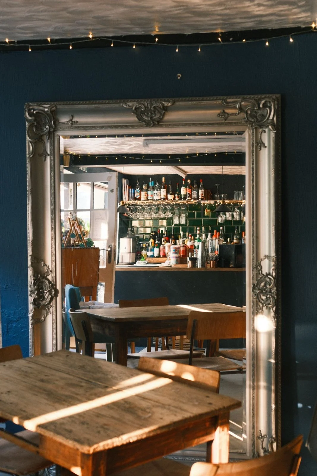 A cozy bar inside a restaurant, seen through a large ornate mirror frame. The bar has various bottles of alcohol and bar accessories, with hanging glasses above. In the foreground, there are wooden tables and chairs, with sunlight casting shadows.