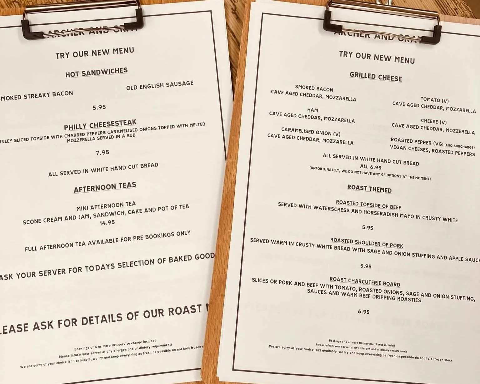 Two menus on clipboards placed on a wooden table, one for hot sandwiches and the other for grilled cheese options, including various types of bacon, ham, cheeses, and roast meats.