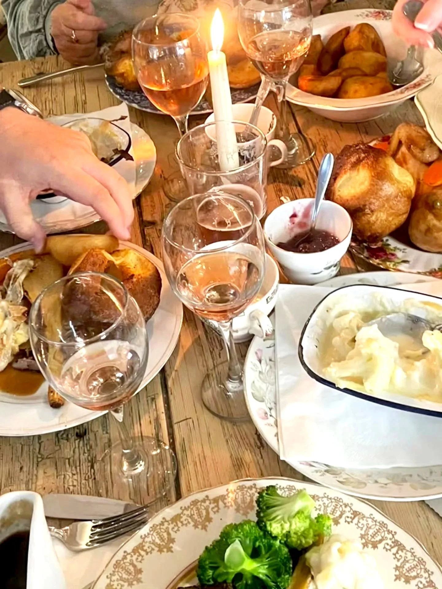 A table set with various dishes, glasses of rosé wine, a lit candle, and some people reaching for food. There are plates of roasted potatoes, a bowl of mashed potatoes, and green broccoli. The table is rustic wood, and the scene suggests a family or 