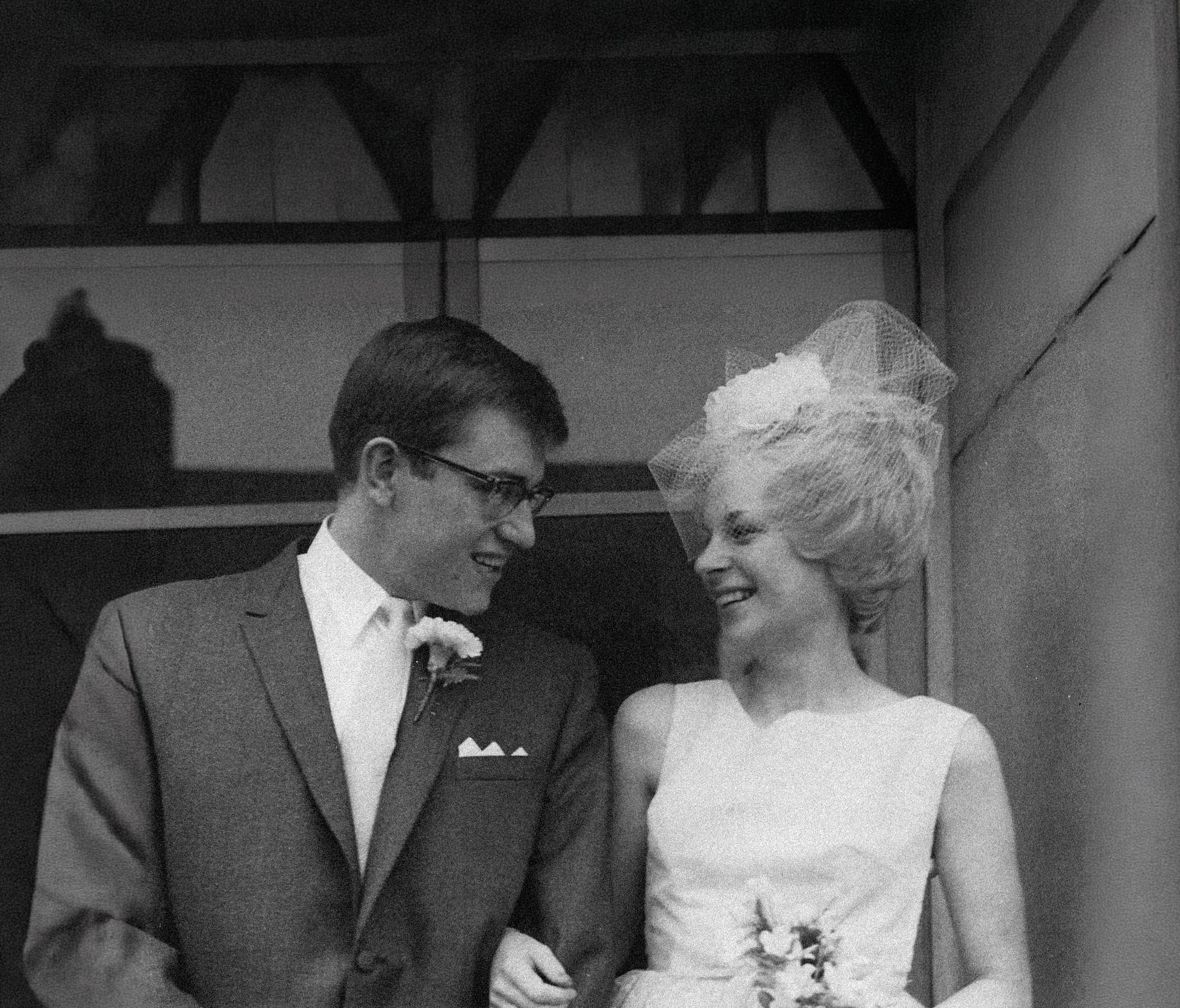 Black and white photo of a man and woman facing each other, smiling, with the woman holding a bouquet of flowers, possibly at a wedding.