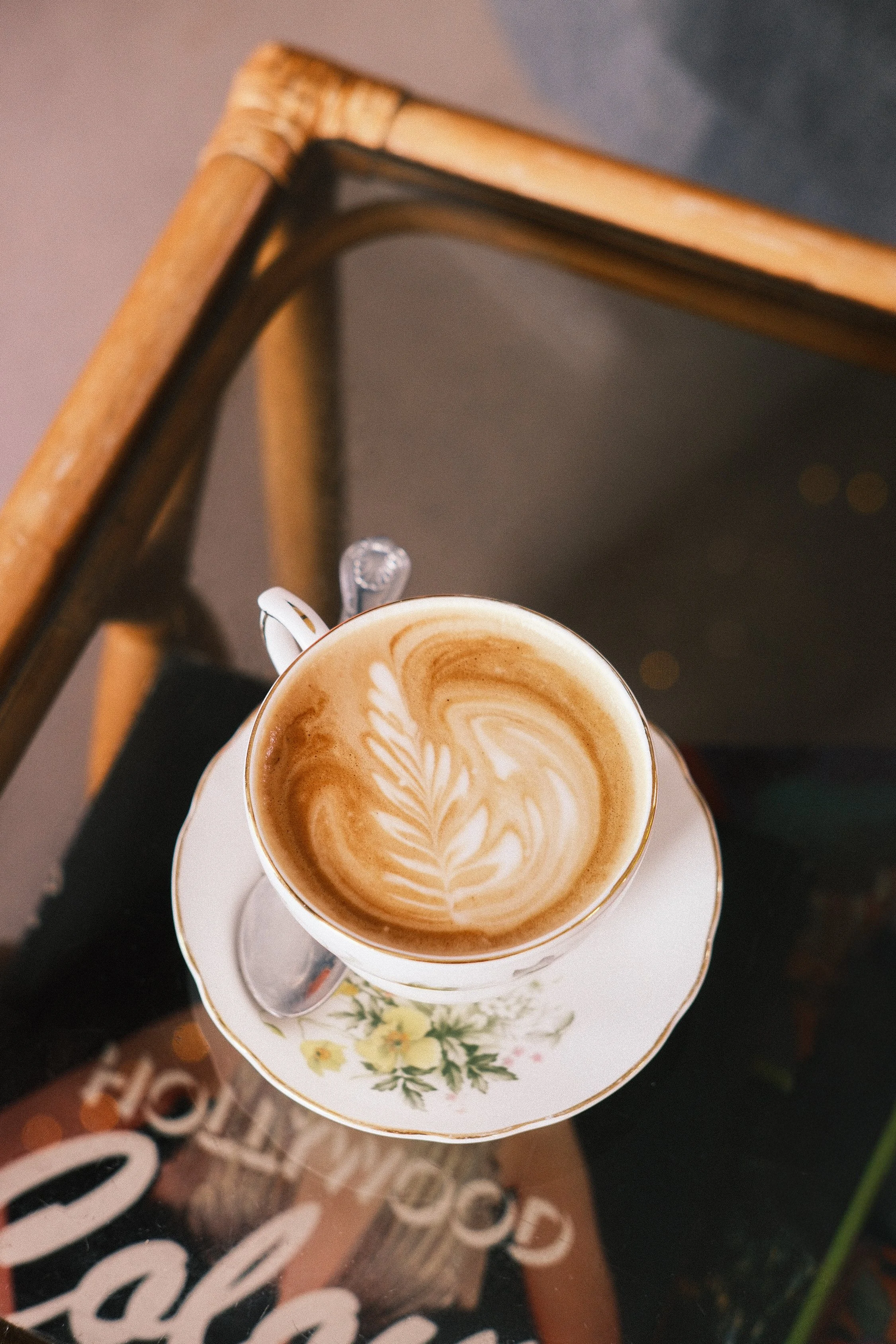 A white porcelain cup with a floral design containing a latte with latte art sitting on a matching saucer, placed on a colorful printed table or surface.