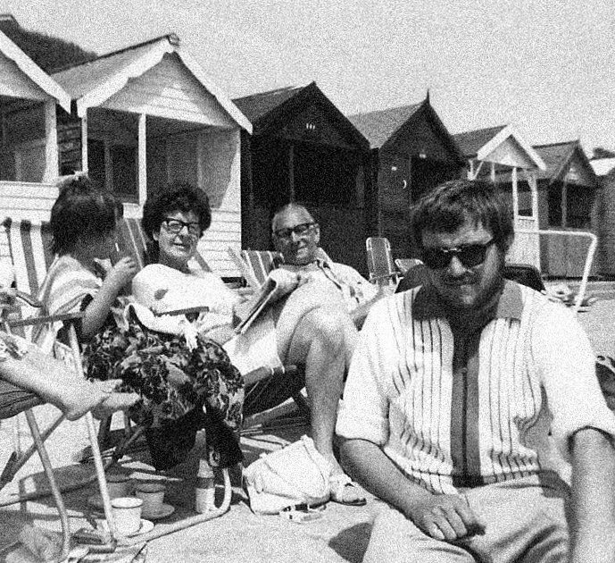A group of four people sitting on beach chairs and relaxing by beach huts, with water in the background, sunny weather, and casual attire.