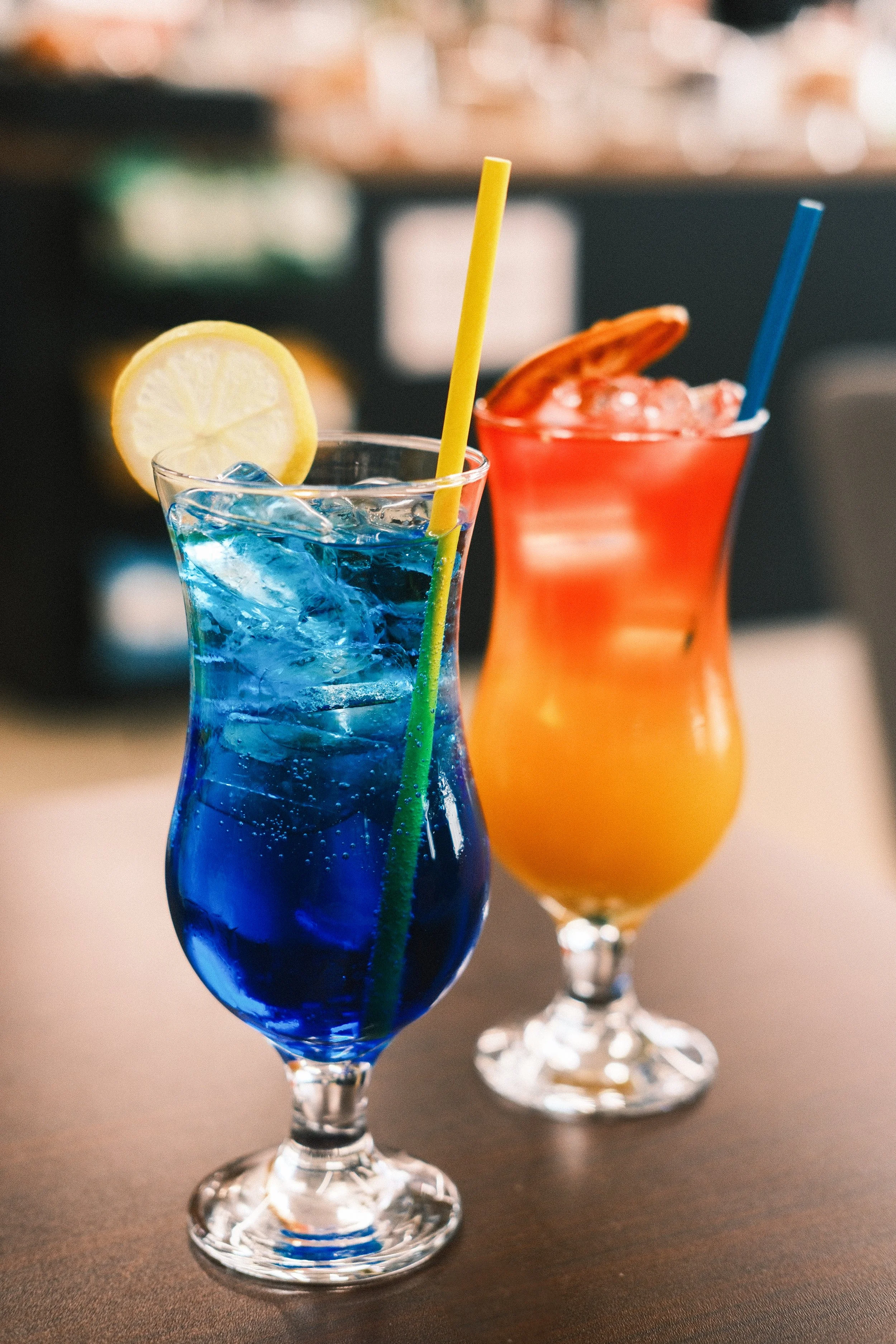 Two tropical cocktails, one blue and one orange, in decorative glassware with straws and garnishes, on a dark table.