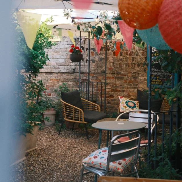 Outdoor patio decorated with colorful bunting banners and paper lanterns, with a wicker chair, a small table, and cushioned chairs, all against a brick wall and surrounded by plants.
