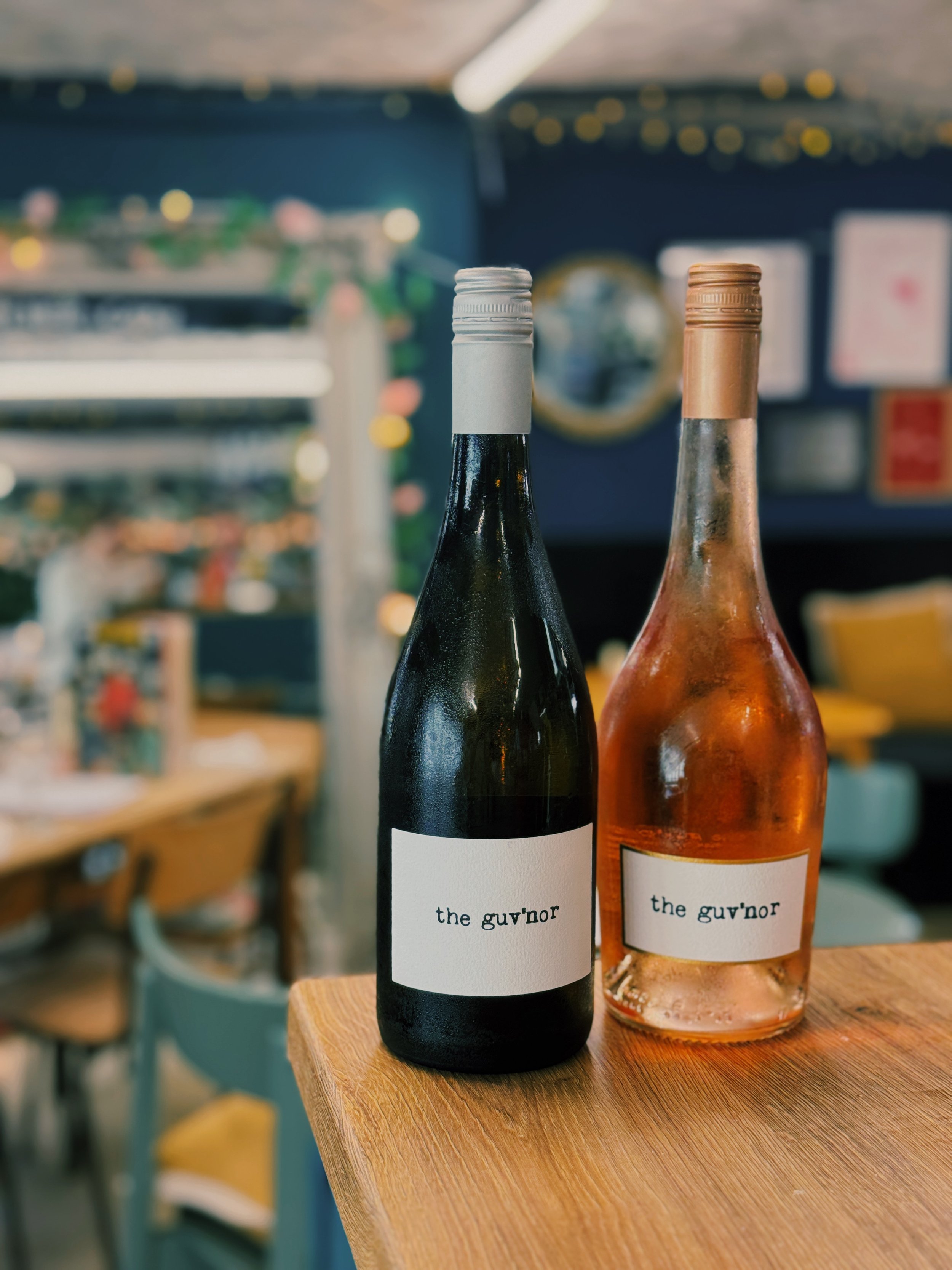 Two wine bottles with label reading 'the guv'nor' placed on a wooden table, with a blurred background of a room decorated with string lights and framed pictures.