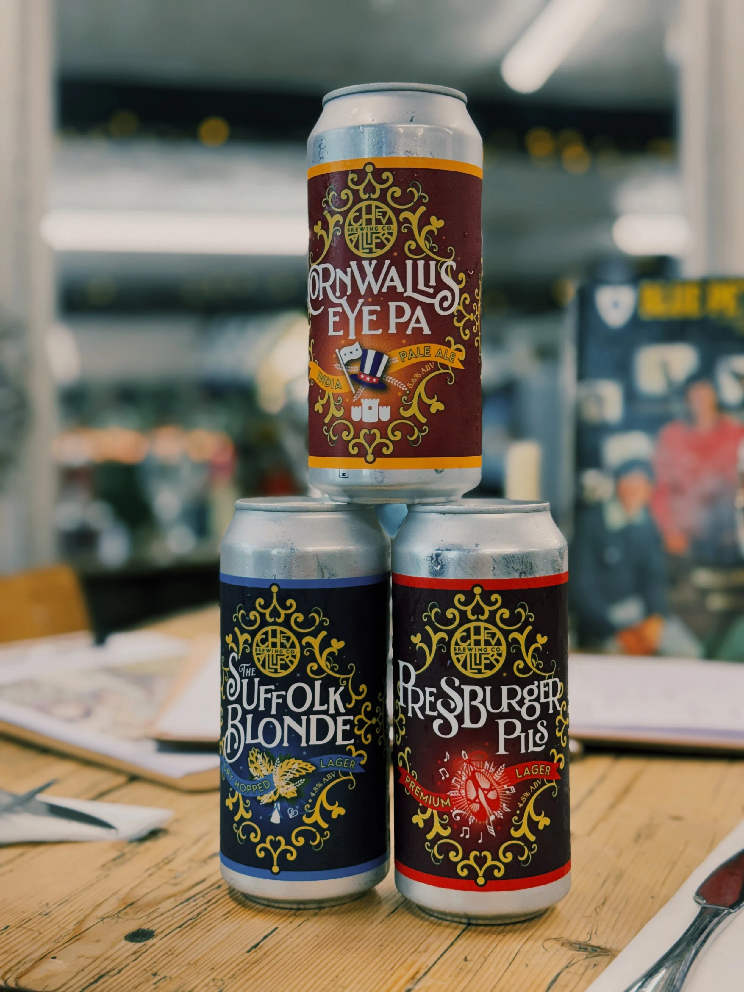Cans of Cornwalls Eye Pale Ale, The Suffolk Blonde Blonde Lager, and Resburger Pils Lager stacked on a wooden table with a blurred background.