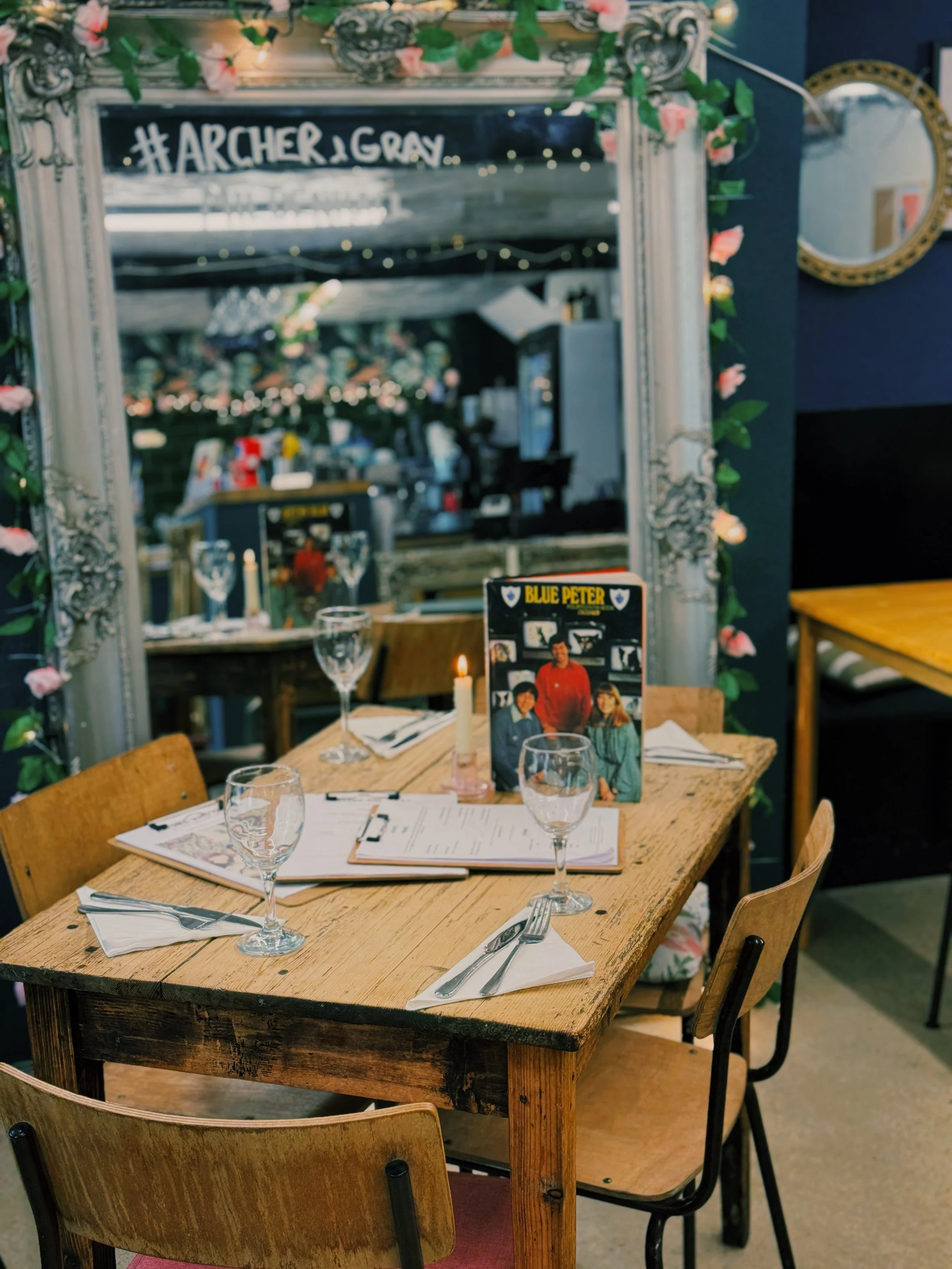A wooden dining table set for four, with menus, napkins, and glasses, in a restaurant decorated with a large mirror framed with flowers, and a sign for the band Archer & Gray visible in the background.