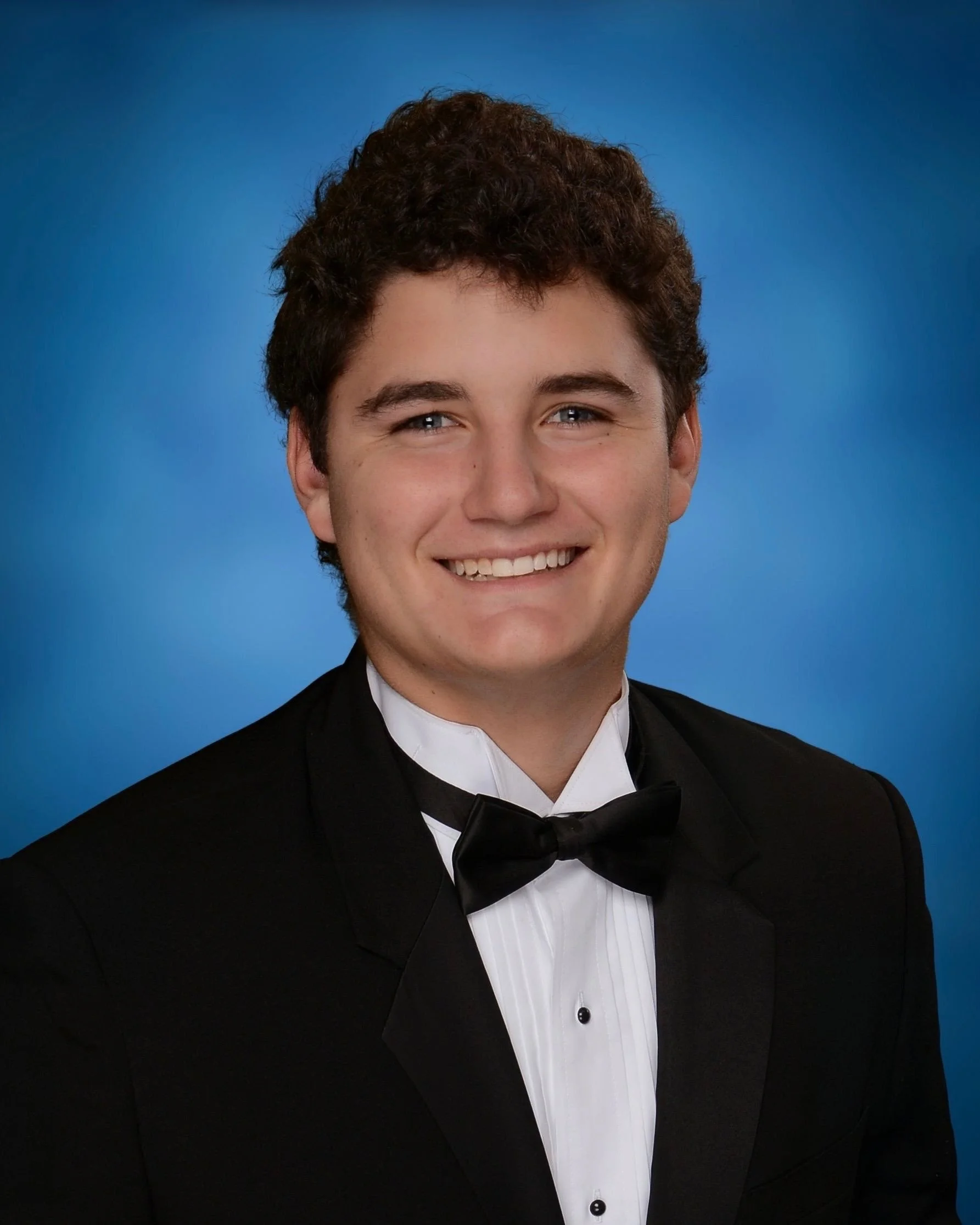 Young man in a tuxedo with a bow tie smiling against a blue background.