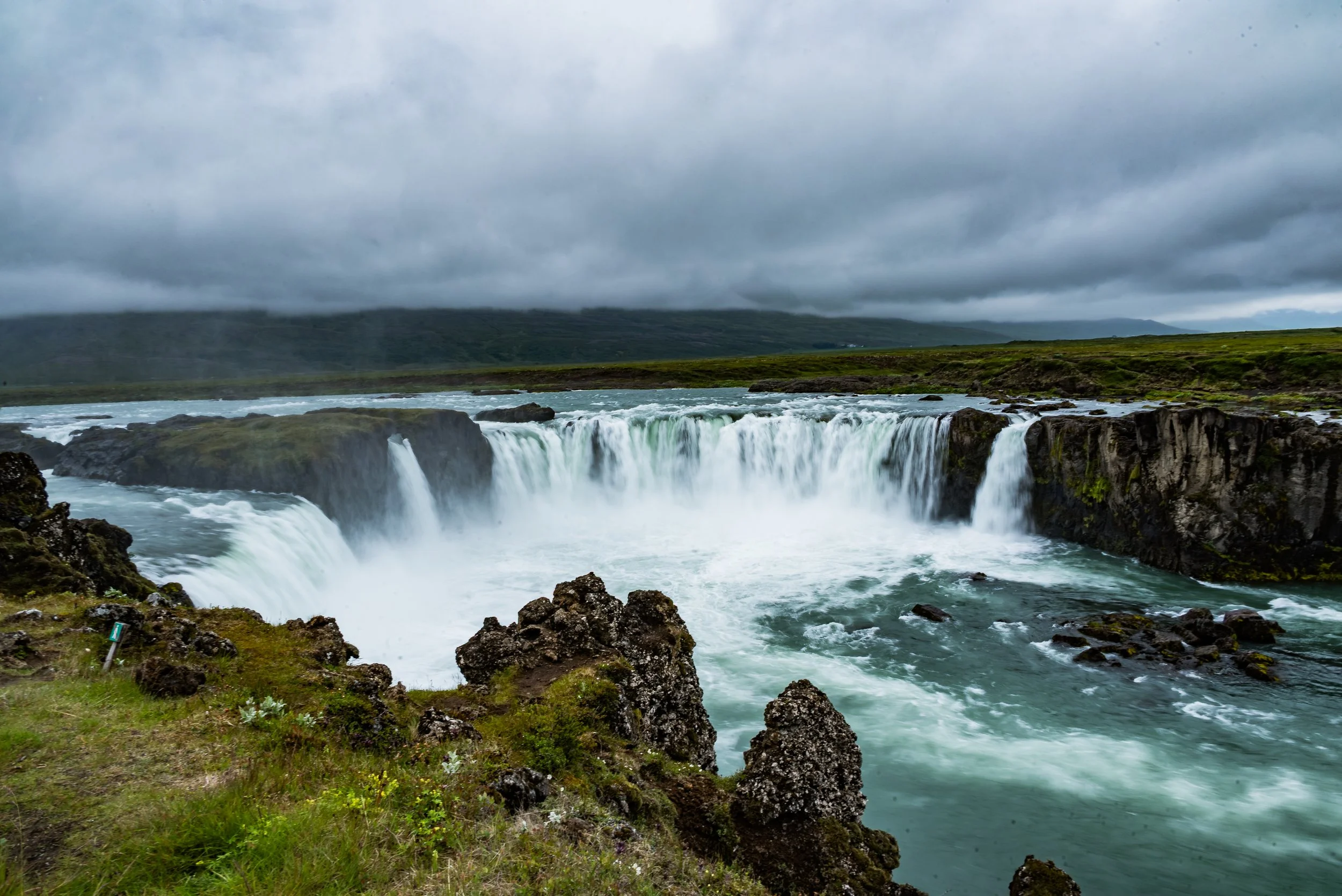 A waterfall cascading over rocks in a cloudy landscape with green hills and a cloudy sky.