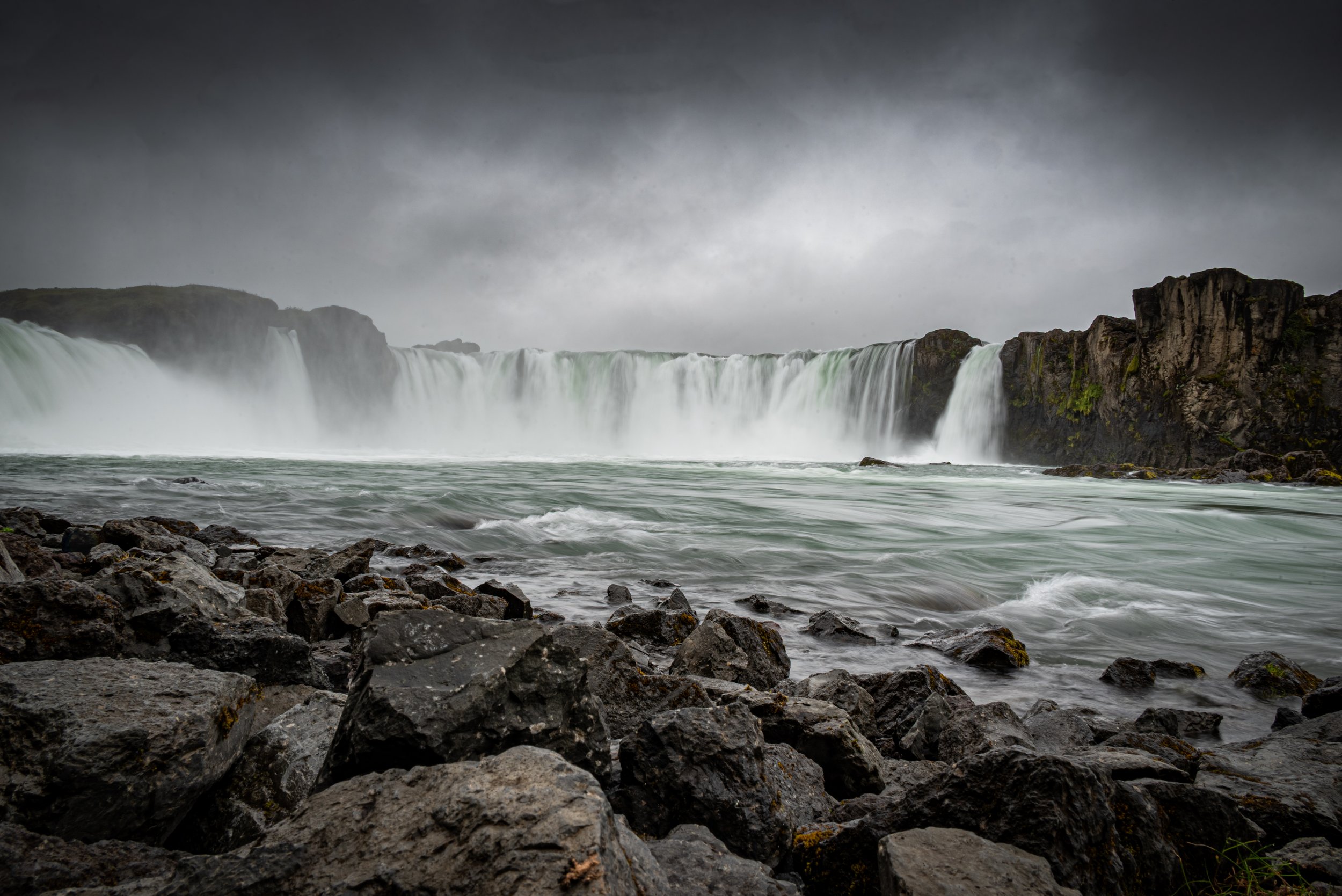 A large waterfall cascading over cliffs with rocky shoreline in the foreground, overcast sky.