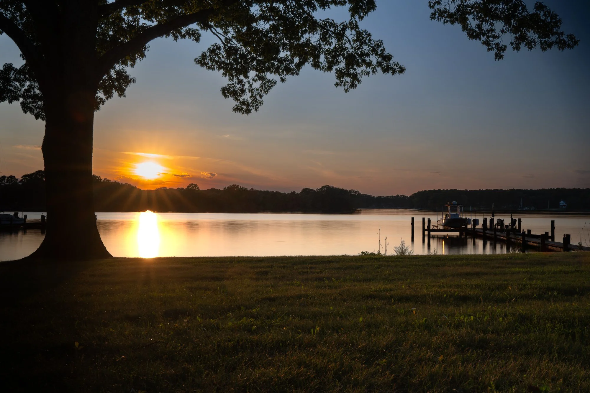 A scenic sunset over a river with a large tree in the foreground and a dock with boats along the water's edge.