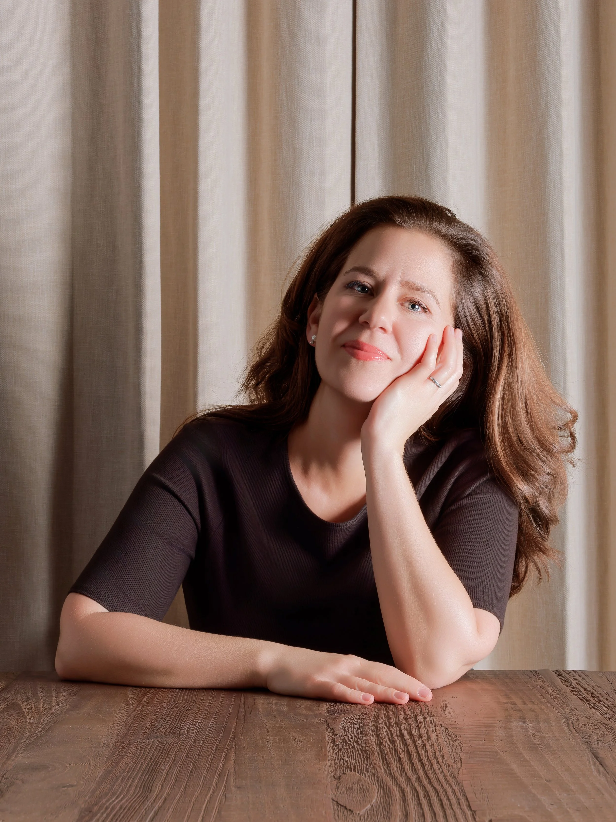 A woman with long wavy brown hair, wearing a dark long-sleeve top, is sitting at a wooden table with her elbow resting on the surface, and her hand resting against her cheek. She is looking directly at the camera with a slight smile, and is positioned in front of beige curtains.